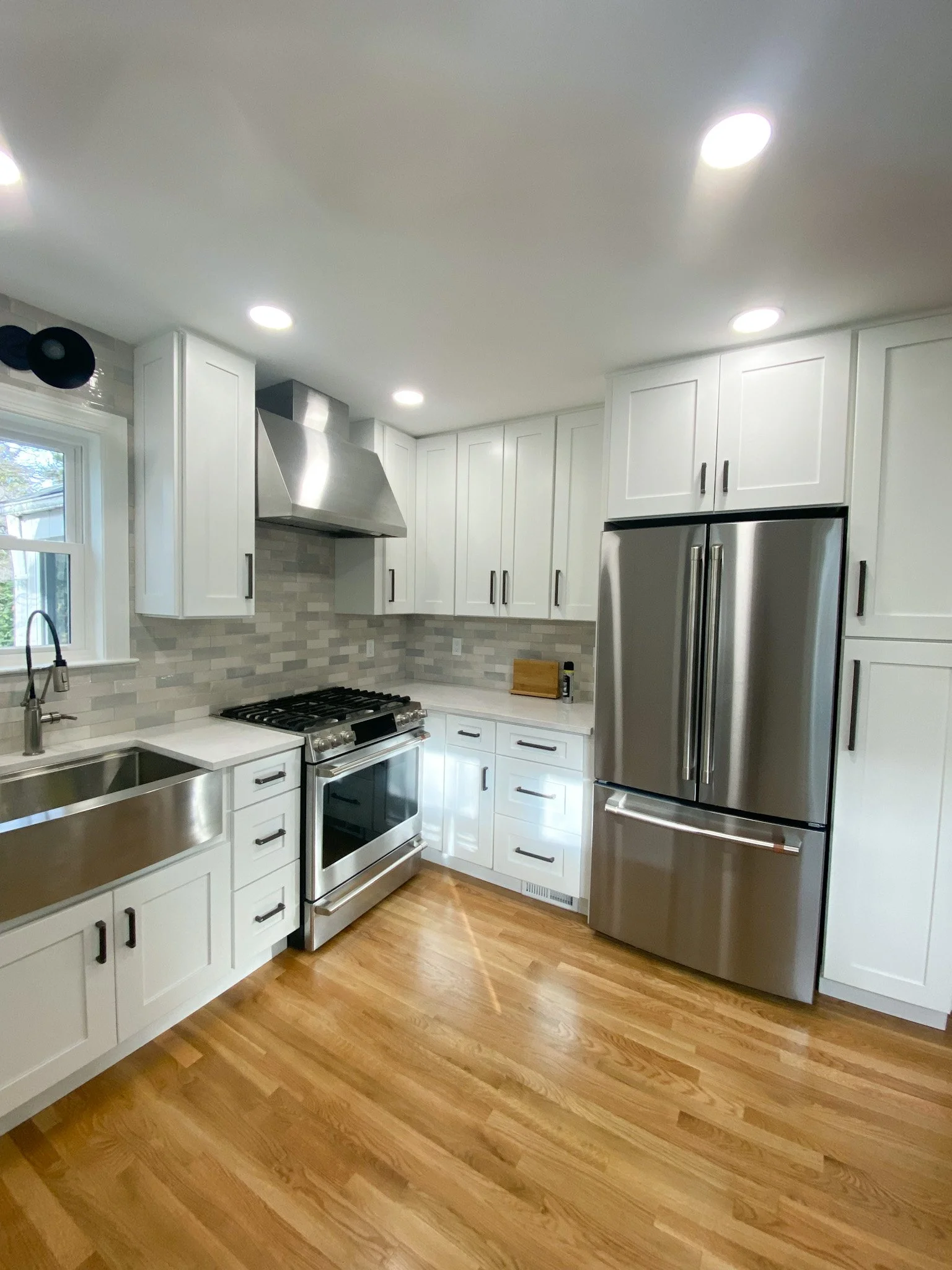 Modern kitchen with white cabinets, stainless steel refrigerator, stainless steel range, gray tile backsplash, hardwood floors, and recessed ceiling lights.