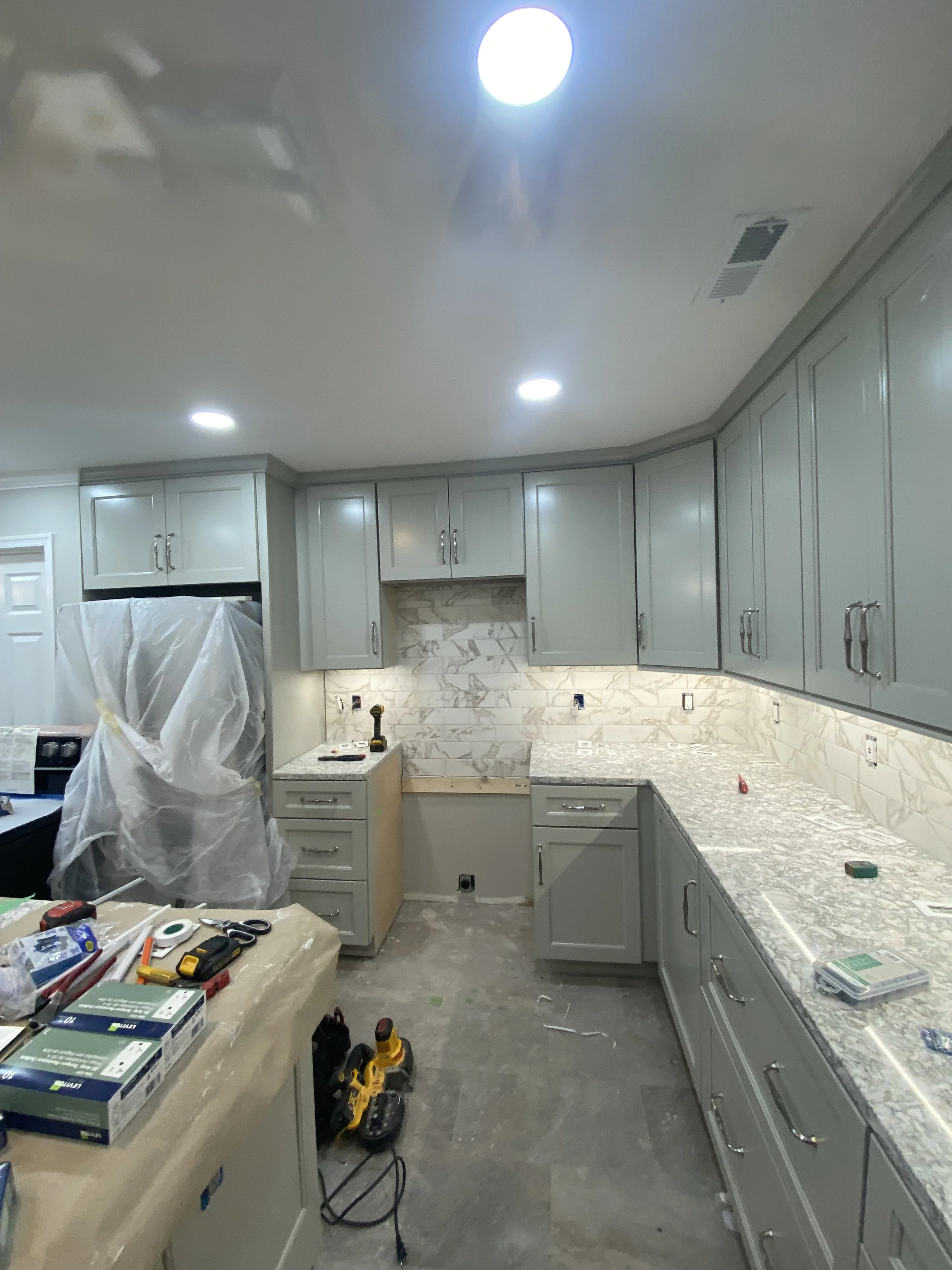 Kitchen under construction with gray cabinets, marble backsplash, and countertops. Construction tools and supplies are on the counter and floor, and a work table with tools is in the foreground.