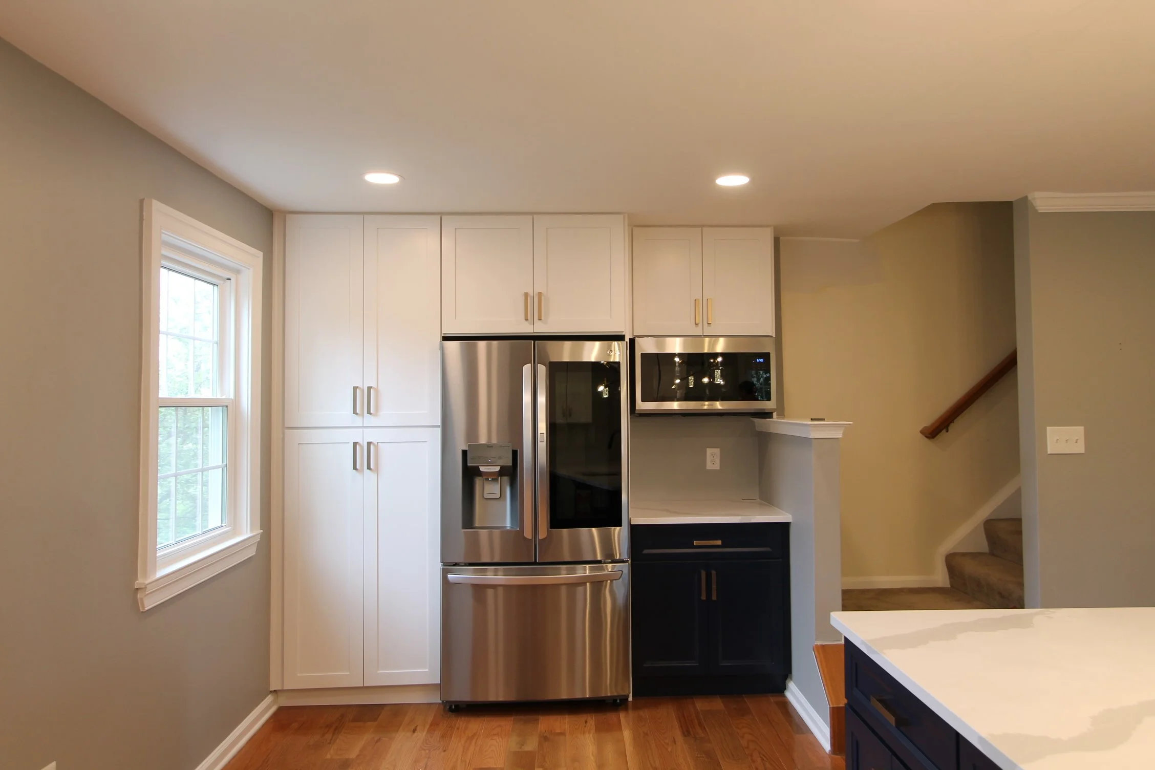 Kitchen with white cabinets, stainless steel refrigerator, microwave, and wooden flooring. There is a window on the left with white trim, and a staircase partially visible on the right.
