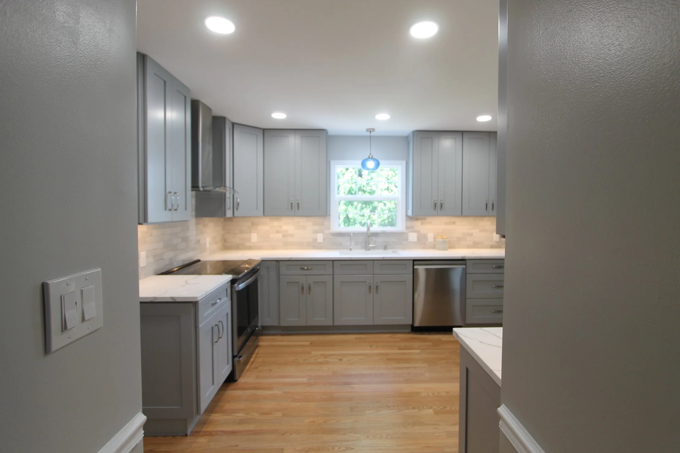 A kitchen with gray cabinets, a window above the sink, a hanging pendant light fixture, stainless steel appliances, hardwood floors, and recessed ceiling lights.