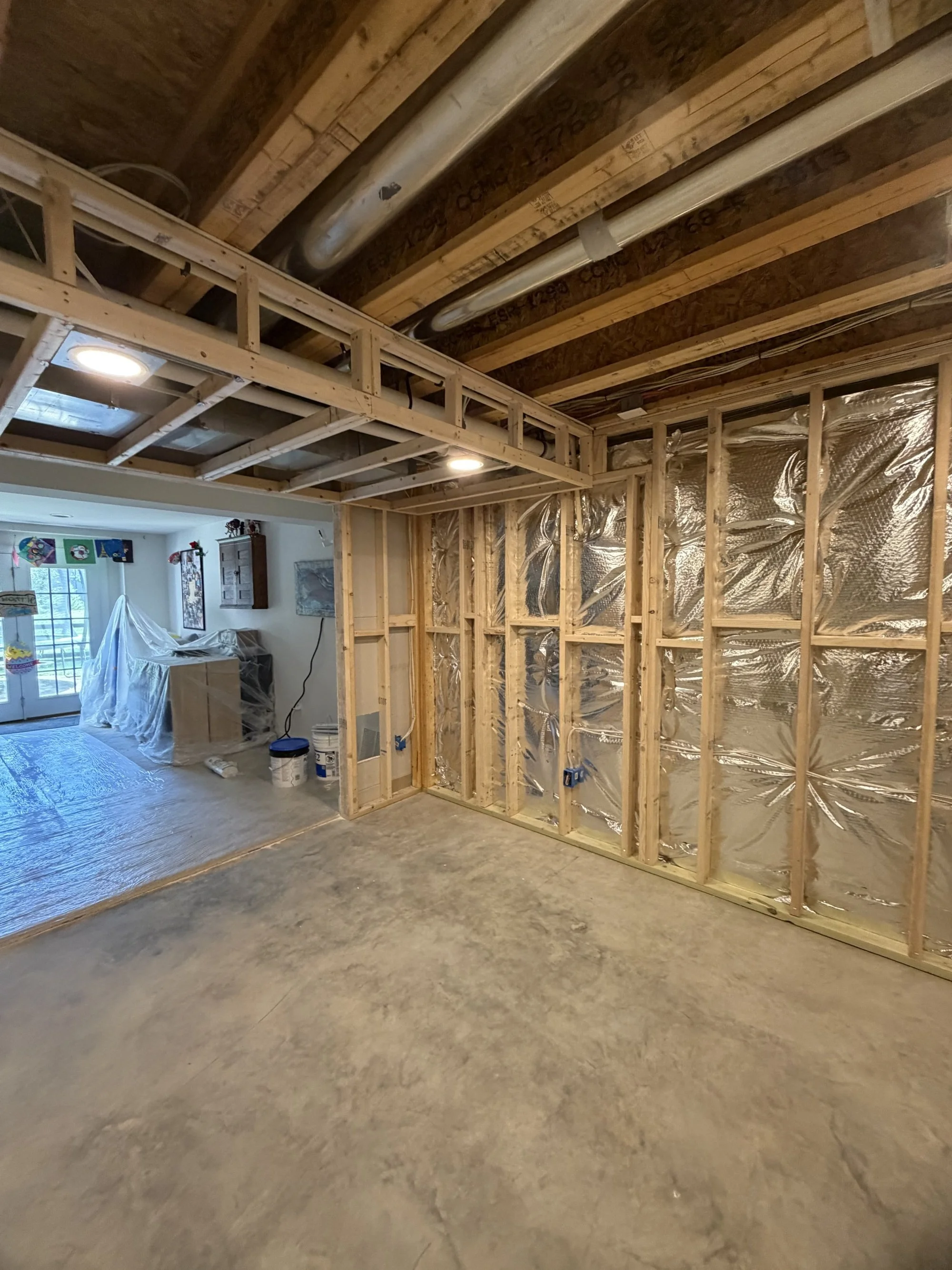 Interior of a house under construction, showing wooden framing, drywall, and unfinished ceiling with visible pipes and insulation.
