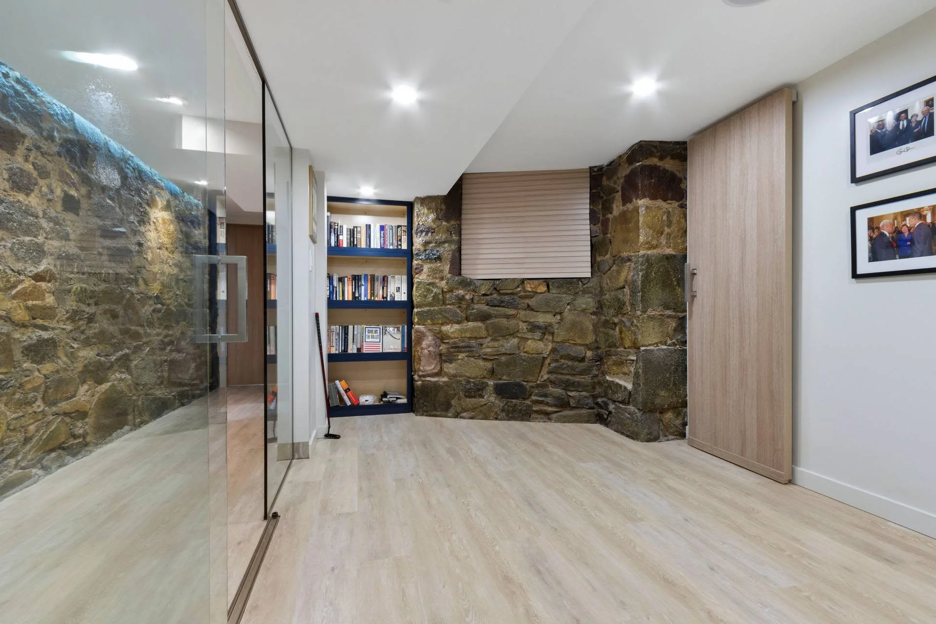 Interior of a modern office or conference room with exposed stone wall, laminate flooring, built-in bookshelf filled with books, glass wall with a metal handle, and framed photos on the white wall.
