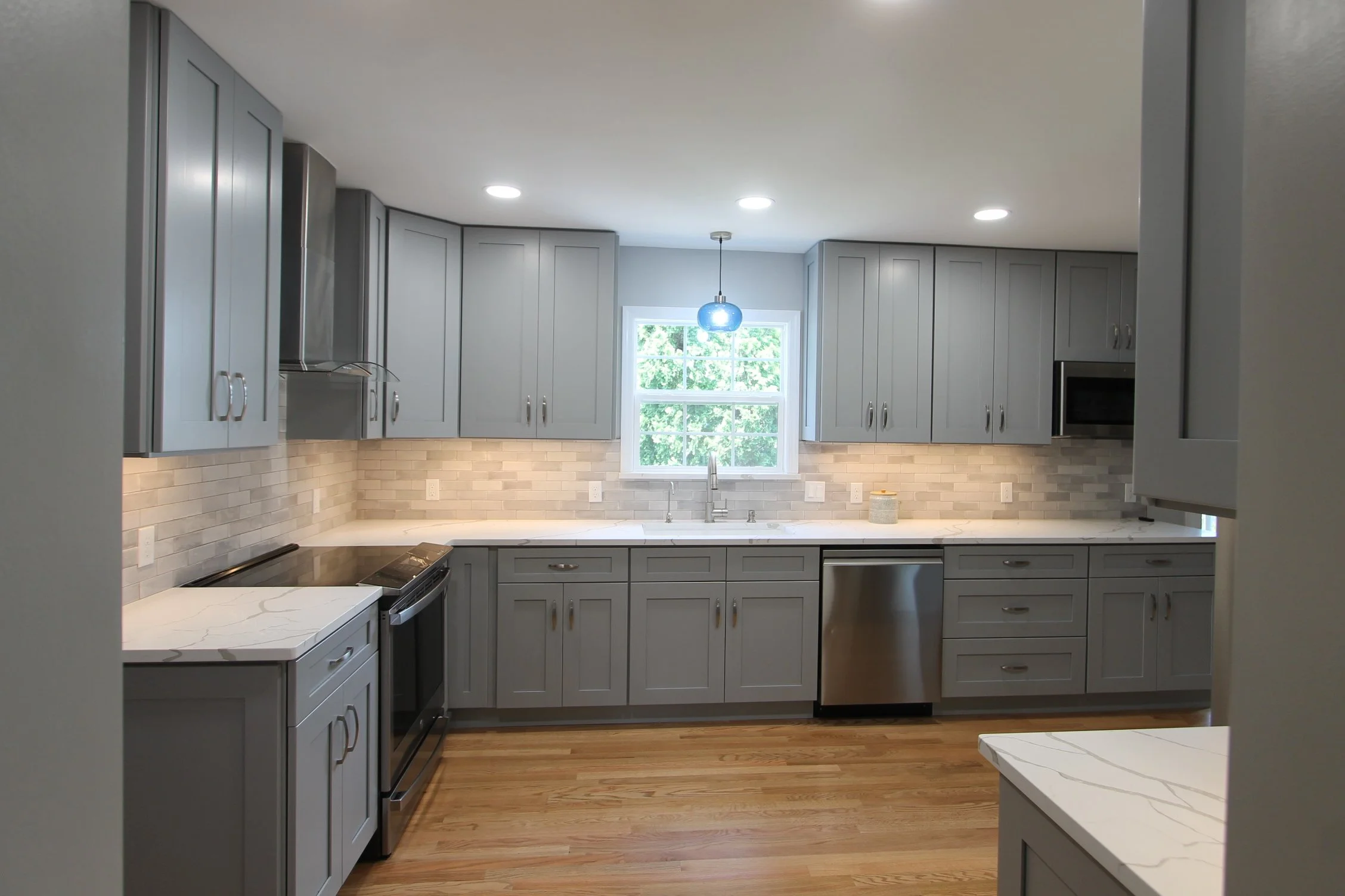 Modern kitchen with gray cabinets, white marble countertops, beige brick backsplash, stainless steel appliances, and a window with greenery outside.