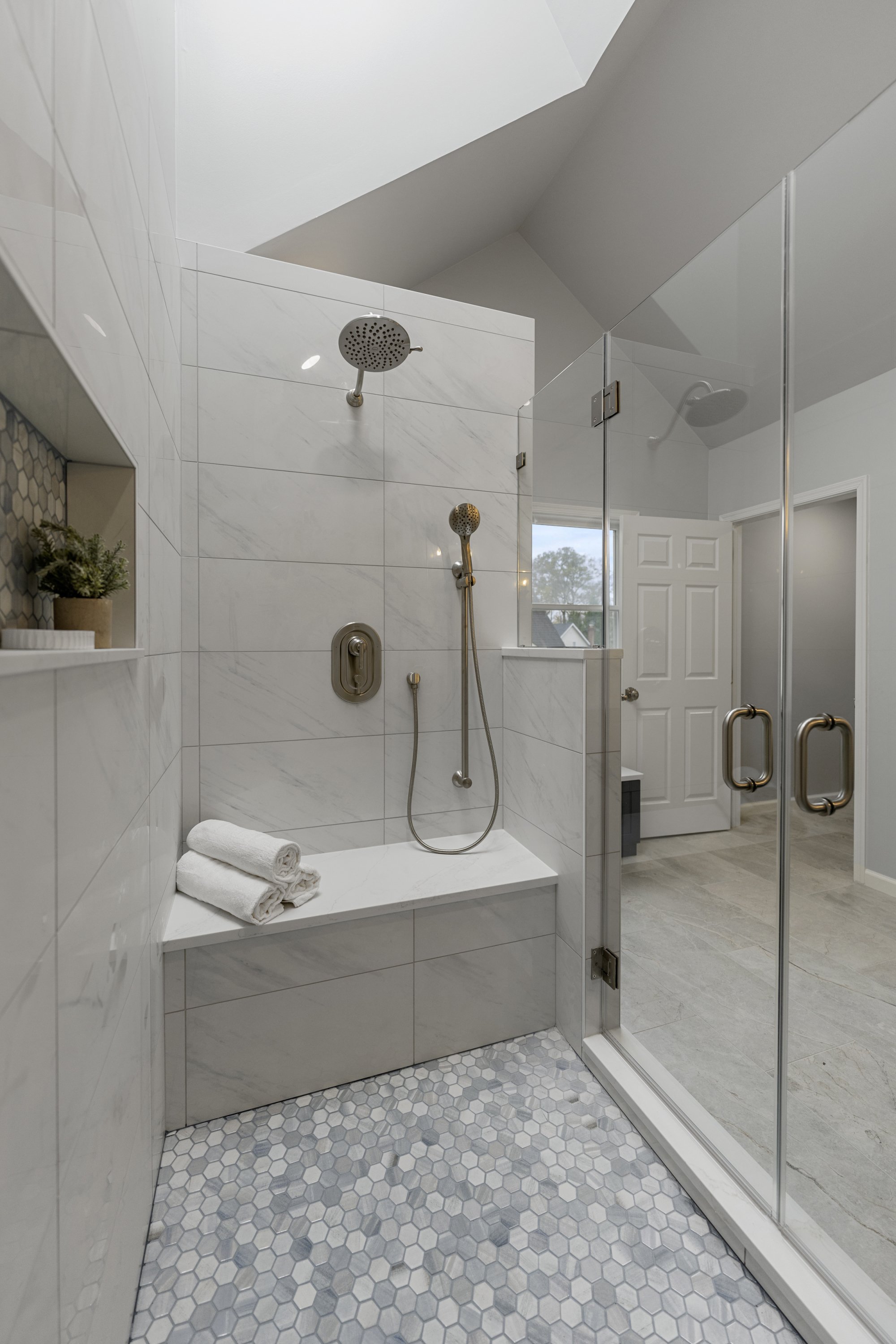 Modern bathroom with a walk-in shower enclosed by glass doors, white hexagonal mosaic tile flooring, and white marble wall tiles. Inside the shower, there are two showerheads, a built-in shelf with a small plant, and folded towels on a bench.