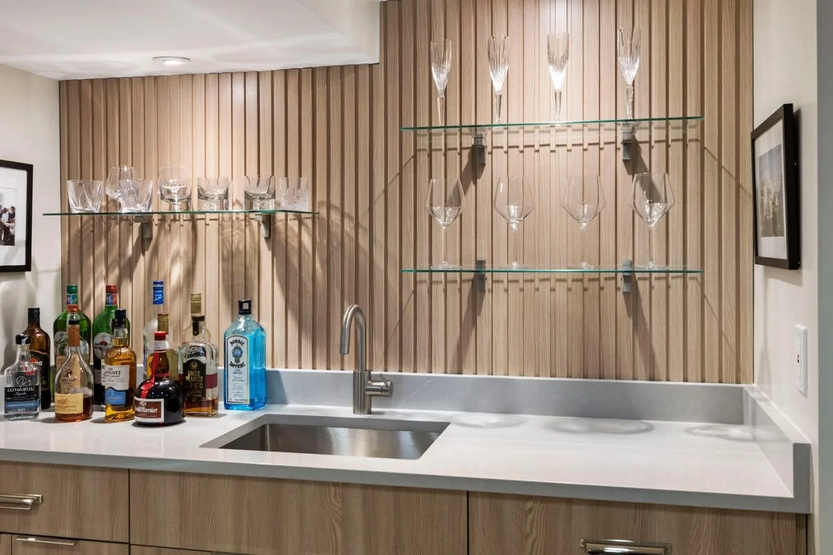 Home bar area with a sink, various bottles of alcohol, and three glass shelves holding empty crystal glasses, set against a wooden panel wall.