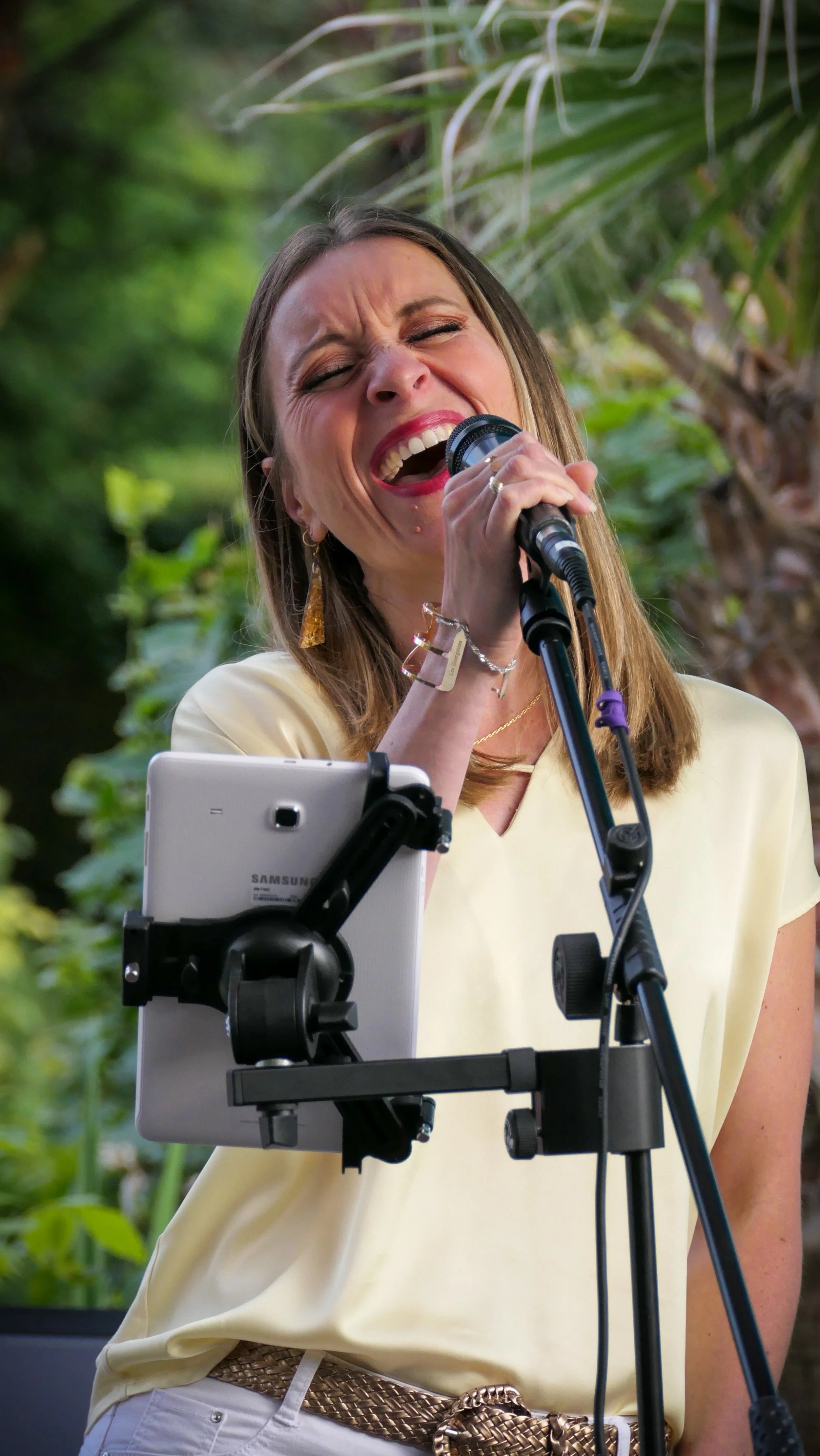 Une femme chante avec passion dans un environnement extérieur vert, utilisant un microphone et une tablette sur un support.