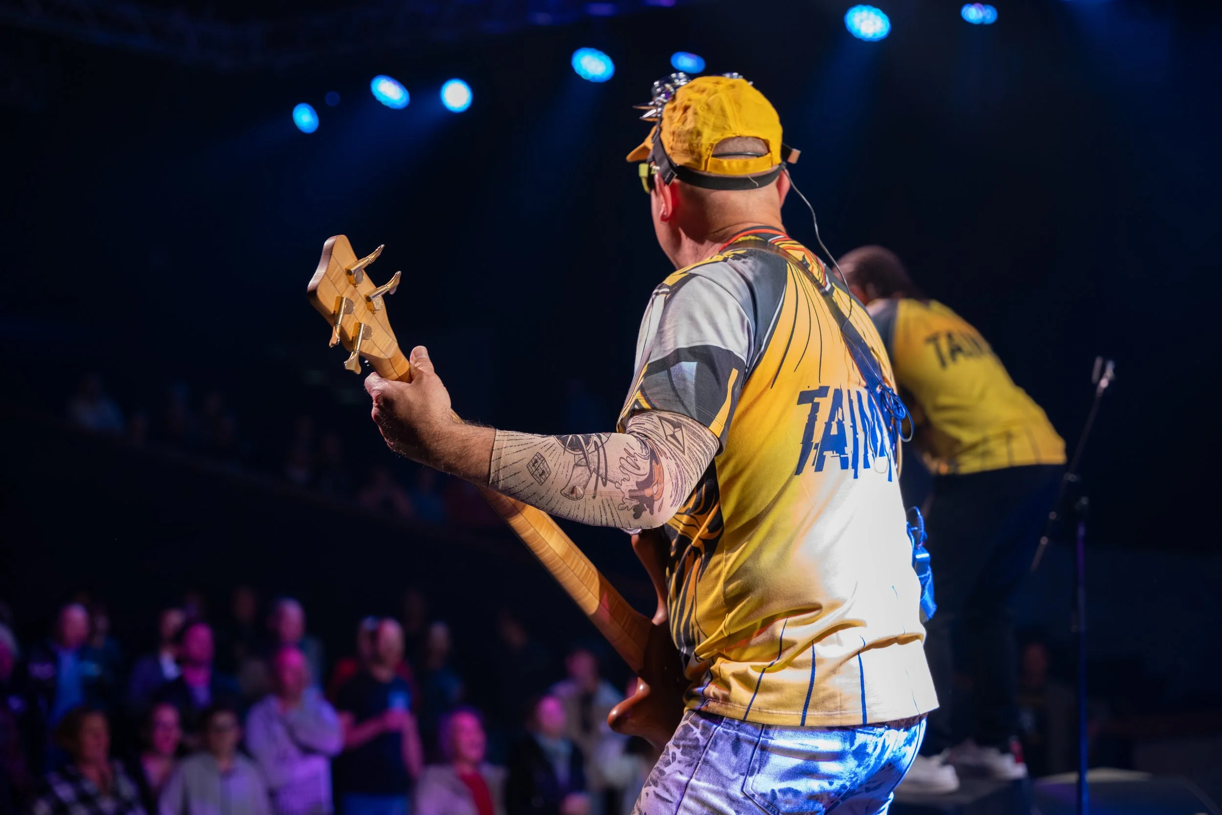 Musicien jouant de la guitare lors d'une performance sur scène, vu de dos. Porte un casque jaune et un t-shirt jaune avec des motifs.