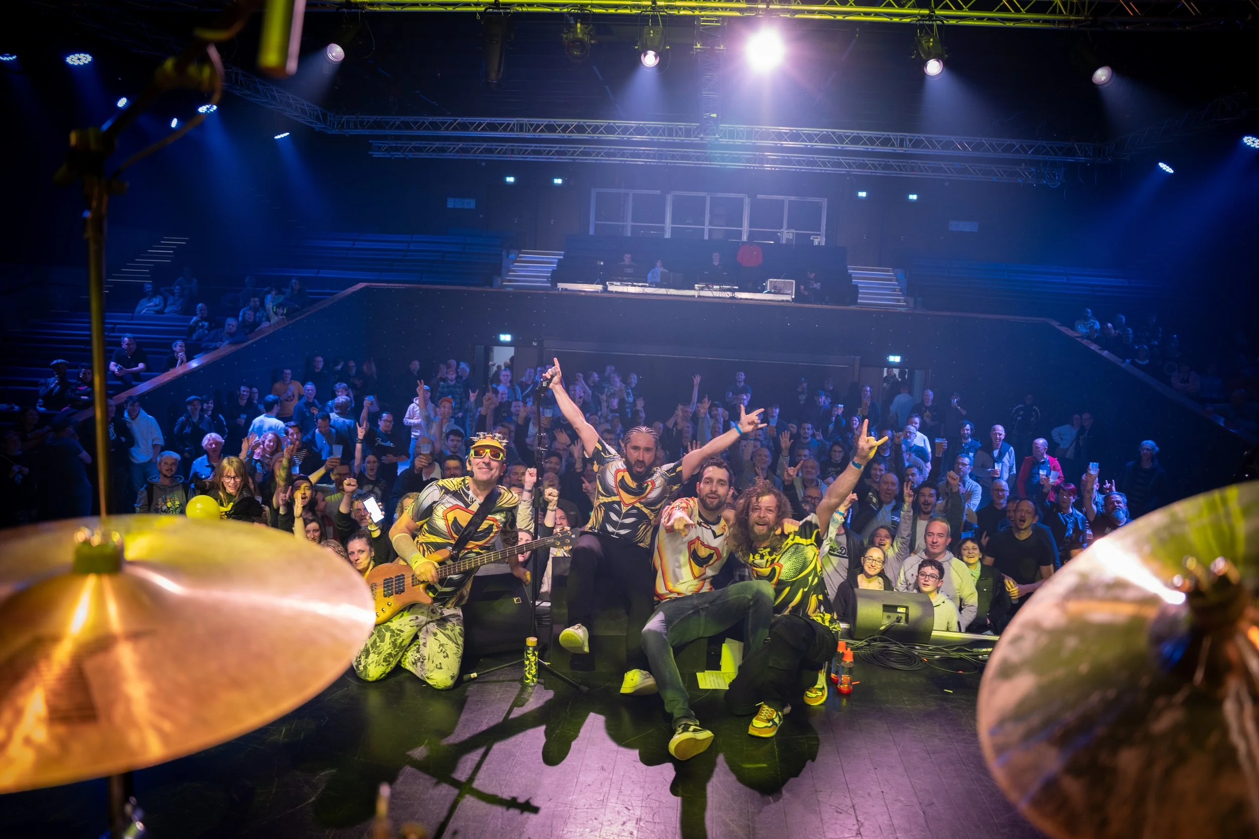 Un groupe de musiciens et de spectateurs dans une salle de concert, vus de la scène avec un drum en premier plan. Les musiciens portent des vêtements colorés et sourient, tandis que le public est enthousiaste et applaudit.