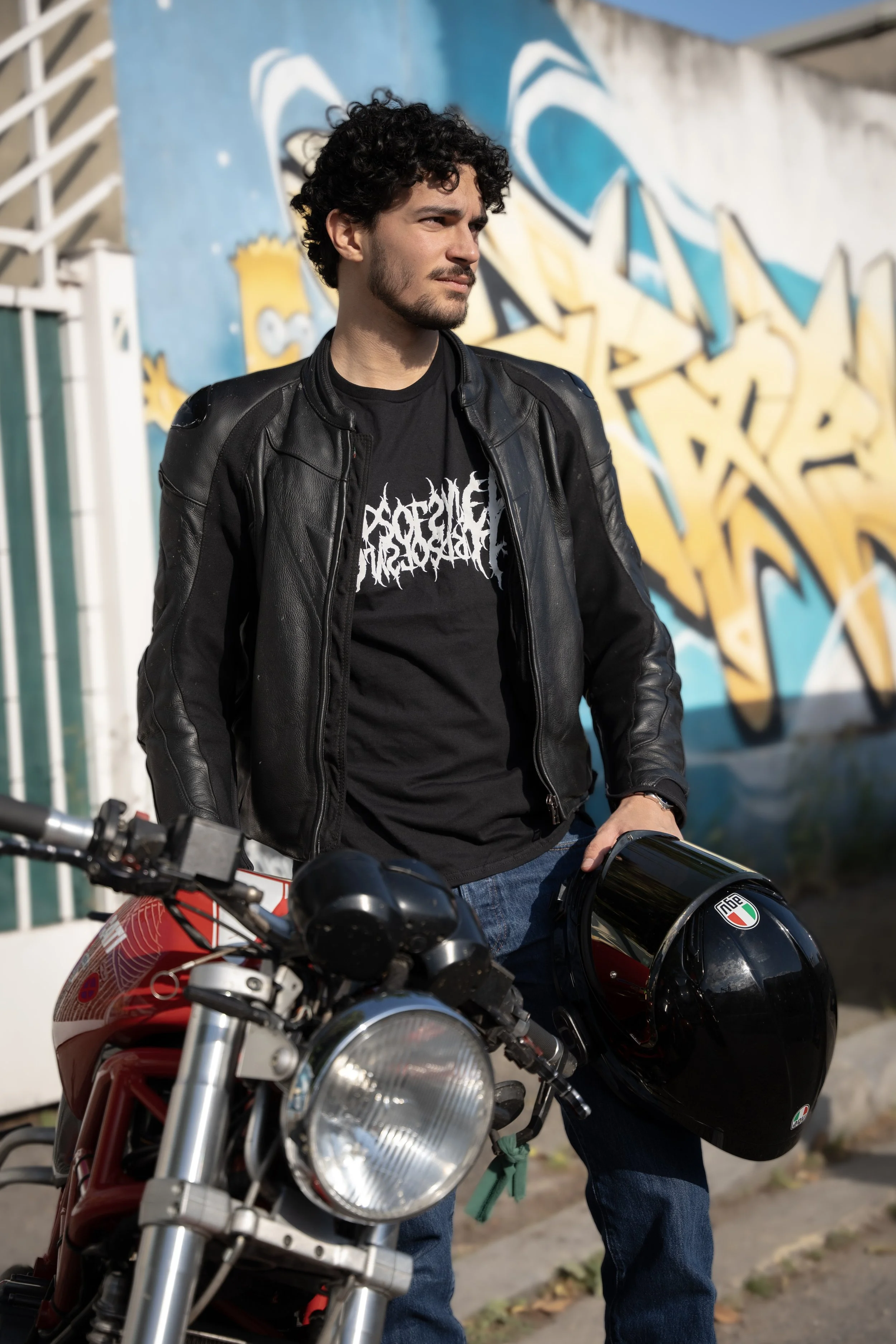 Un jeune homme avec des cheveux bouclés et une barbe, portant une veste en cuir, tient un casque de moto et se trouve à côté d'une moto rouge devant un mur avec des graffitis colorés.