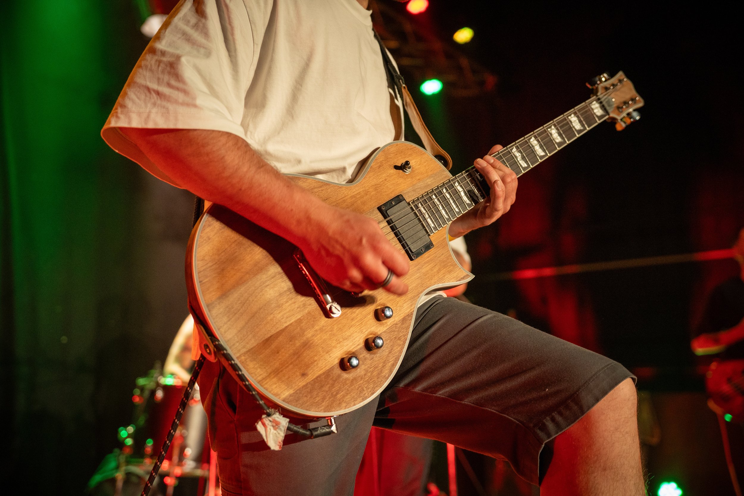 Un musicien joue de la guitare électrique sur scène dans une lumière tamisée avec des lumières vertes et rouges en arrière-plan.