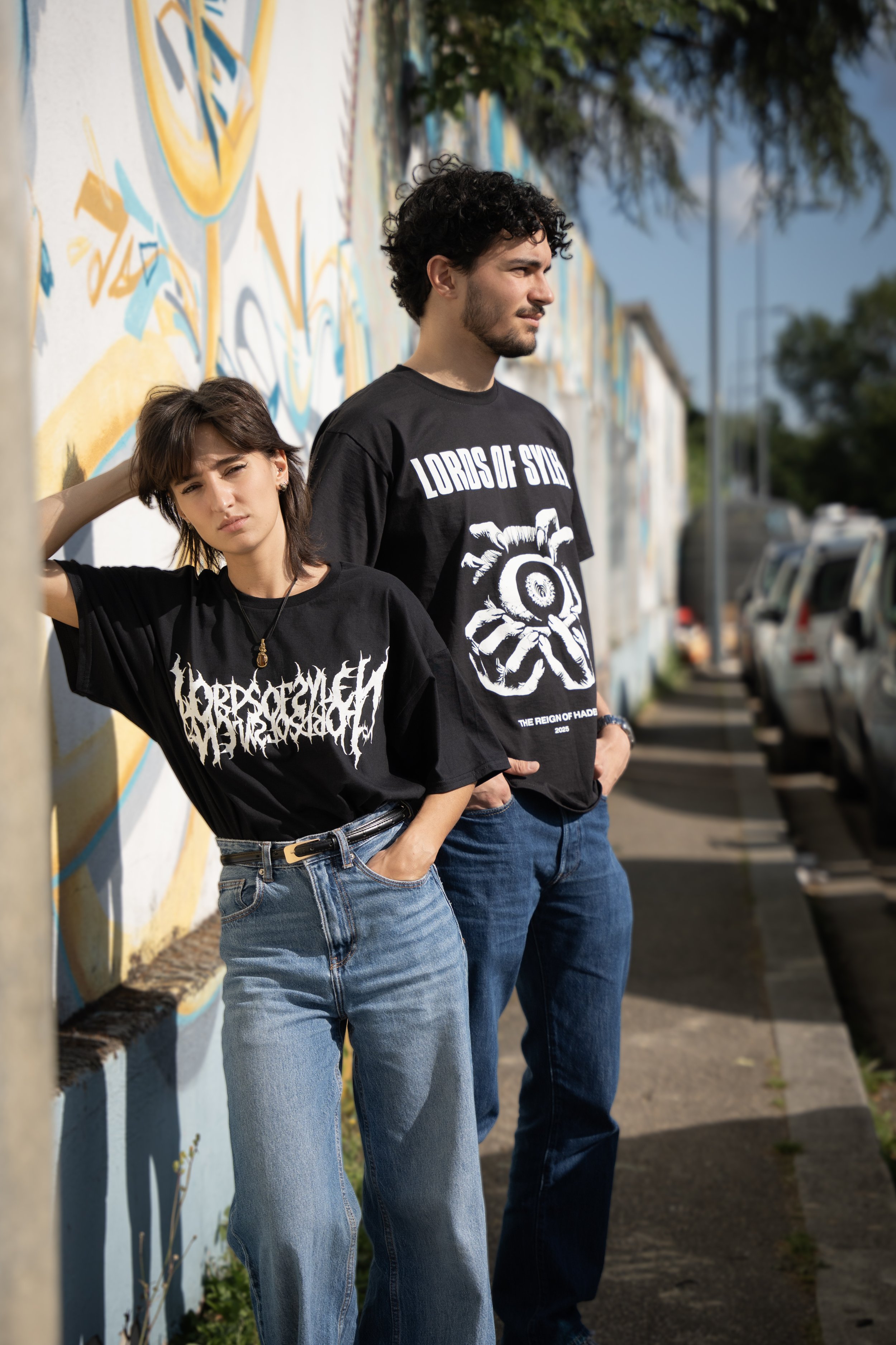 Deux jeunes hommes posent contre un mur graffiti en plein soleil, portant des t-shirts noirs à motifs graphiques, en jeans, dans une rue en arrière-plan avec des voitures stationnées.