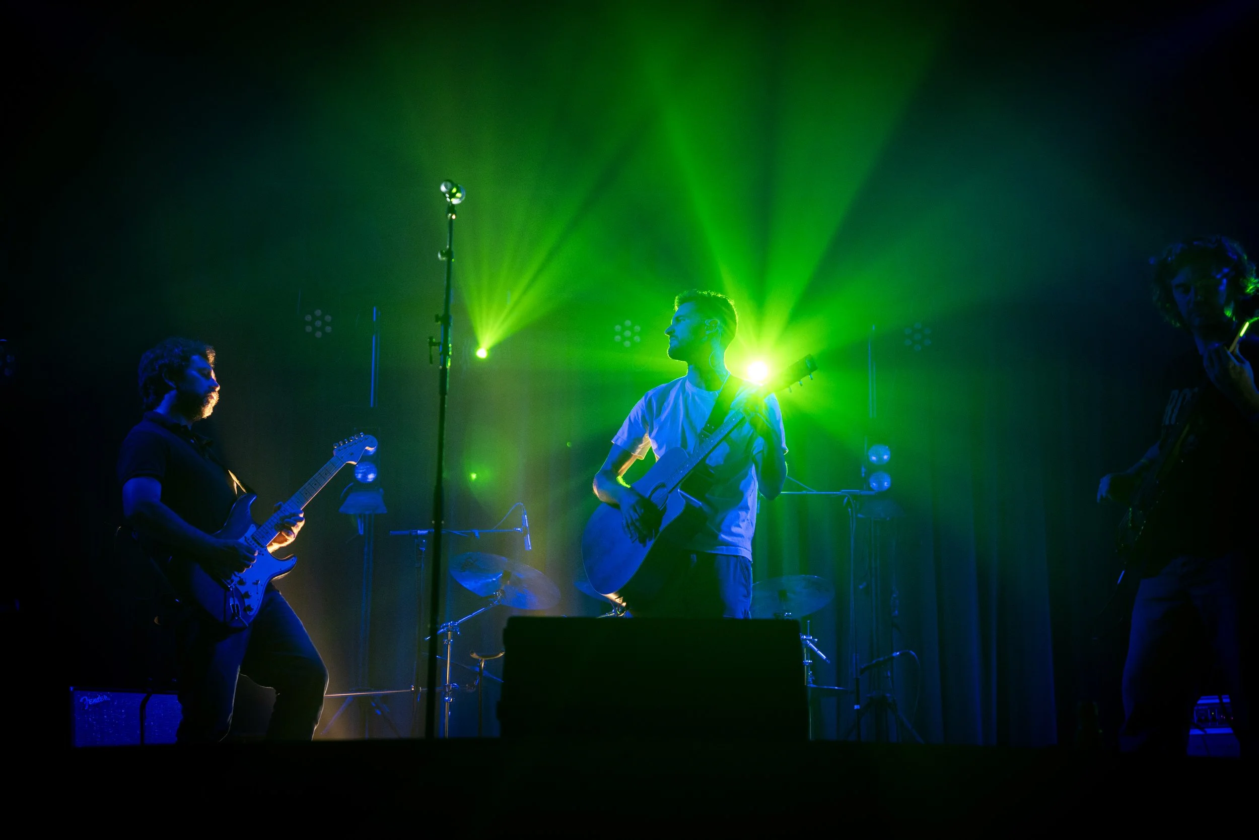 Groupe de musiciens jouant sur scène sous des lumières vertes et bleues, avec un guitariste au centre.