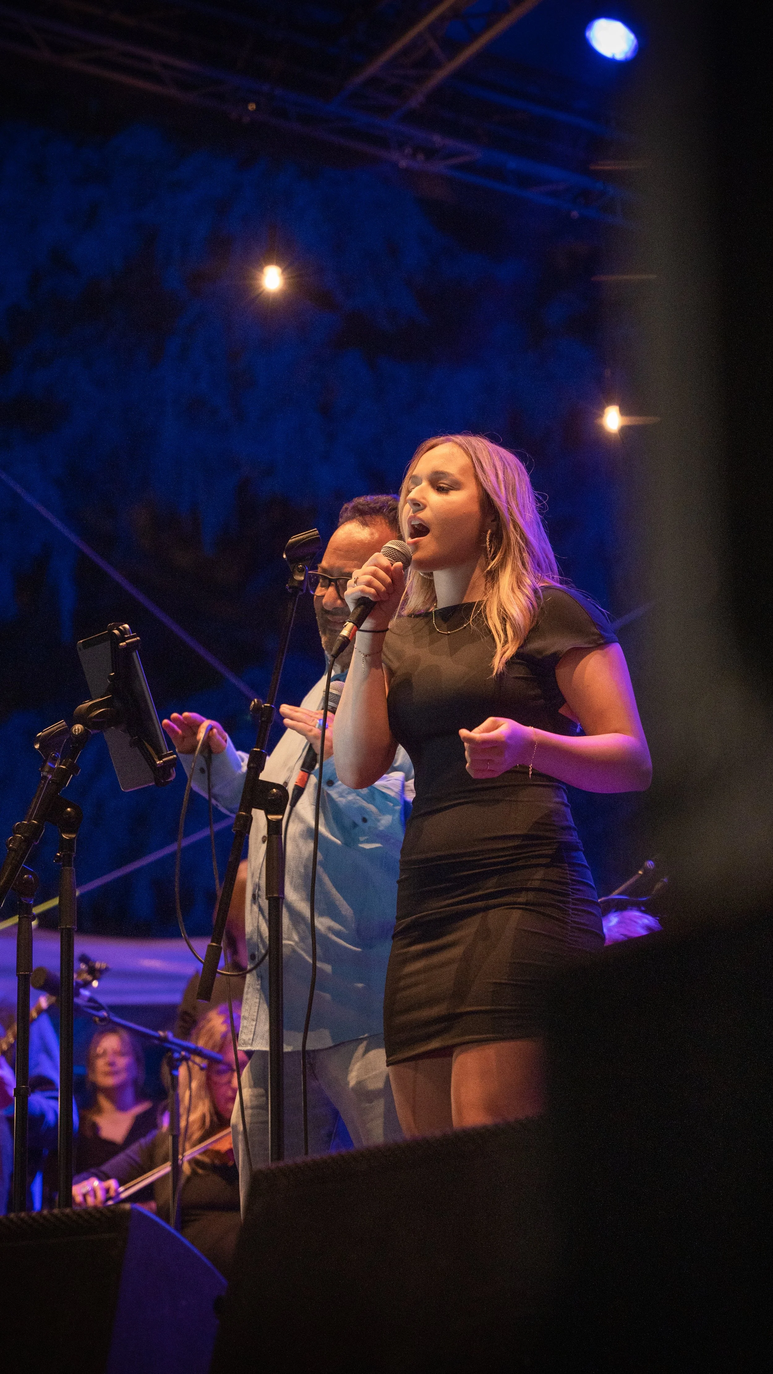 Une femme chante dans un concert avec un microphone, accompagnée d'un homme en arrière-plan, dans une scène sombre avec des éclairages colorés.