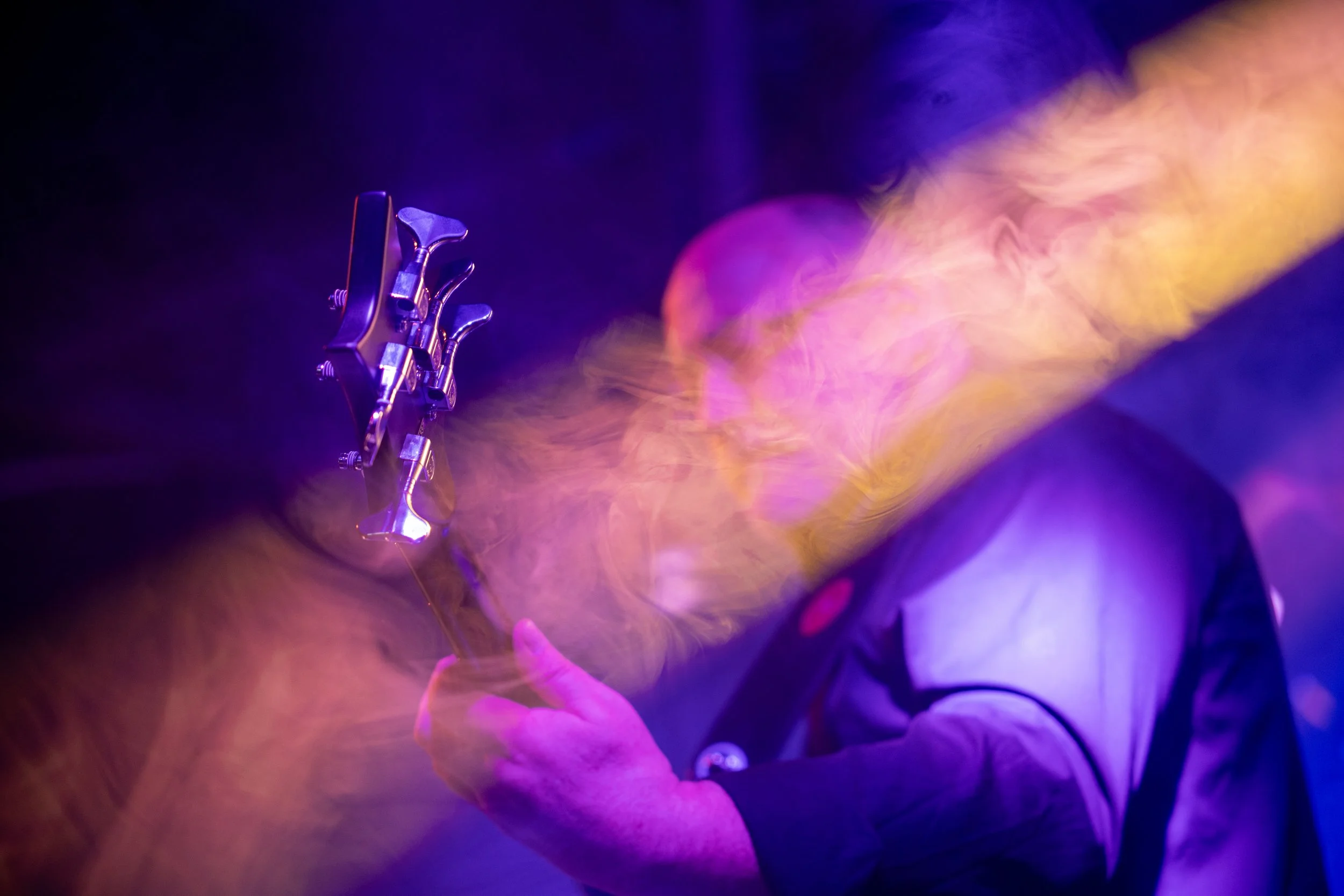 Un guitariste jouant sous un éclairage coloré violet et jaune avec des volutes de fumée ou de vapeur en arrière-plan.