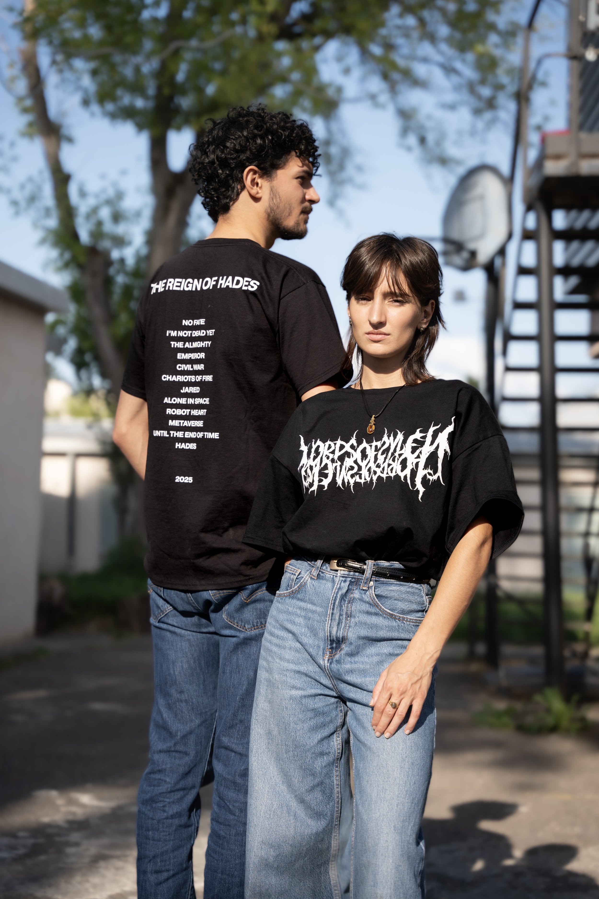 Deux jeunes adultes posent à l'extérieur sous un arbre, portant des t-shirts noirs avec des textes graphiques, un homme a les cheveux noirs bouclés, une femme a les cheveux bruns courts, tous deux en denim.
