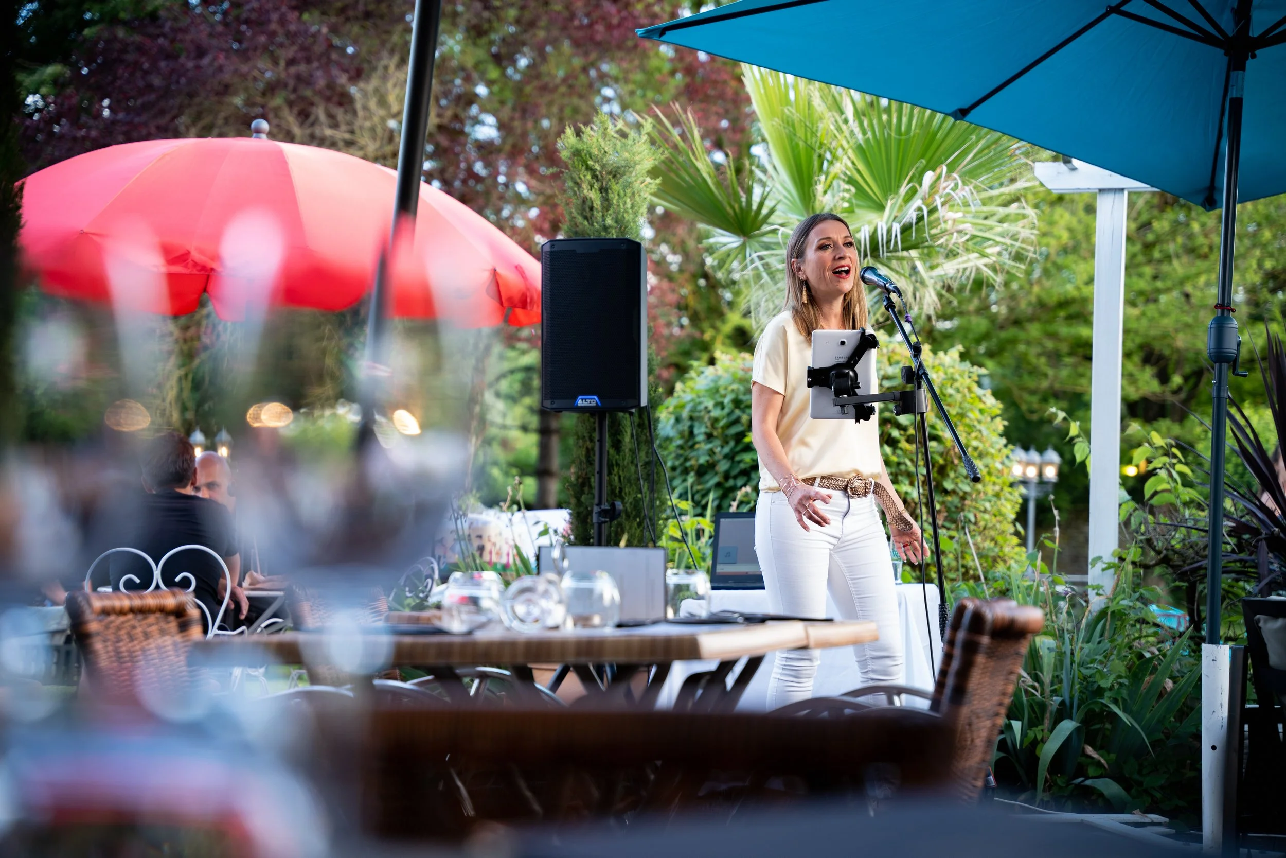 Une femme chante en public lors d'un événement en plein air, sous deux parasols bleus et roses, avec un décor naturel de plantes et d'arbres en arrière-plan.