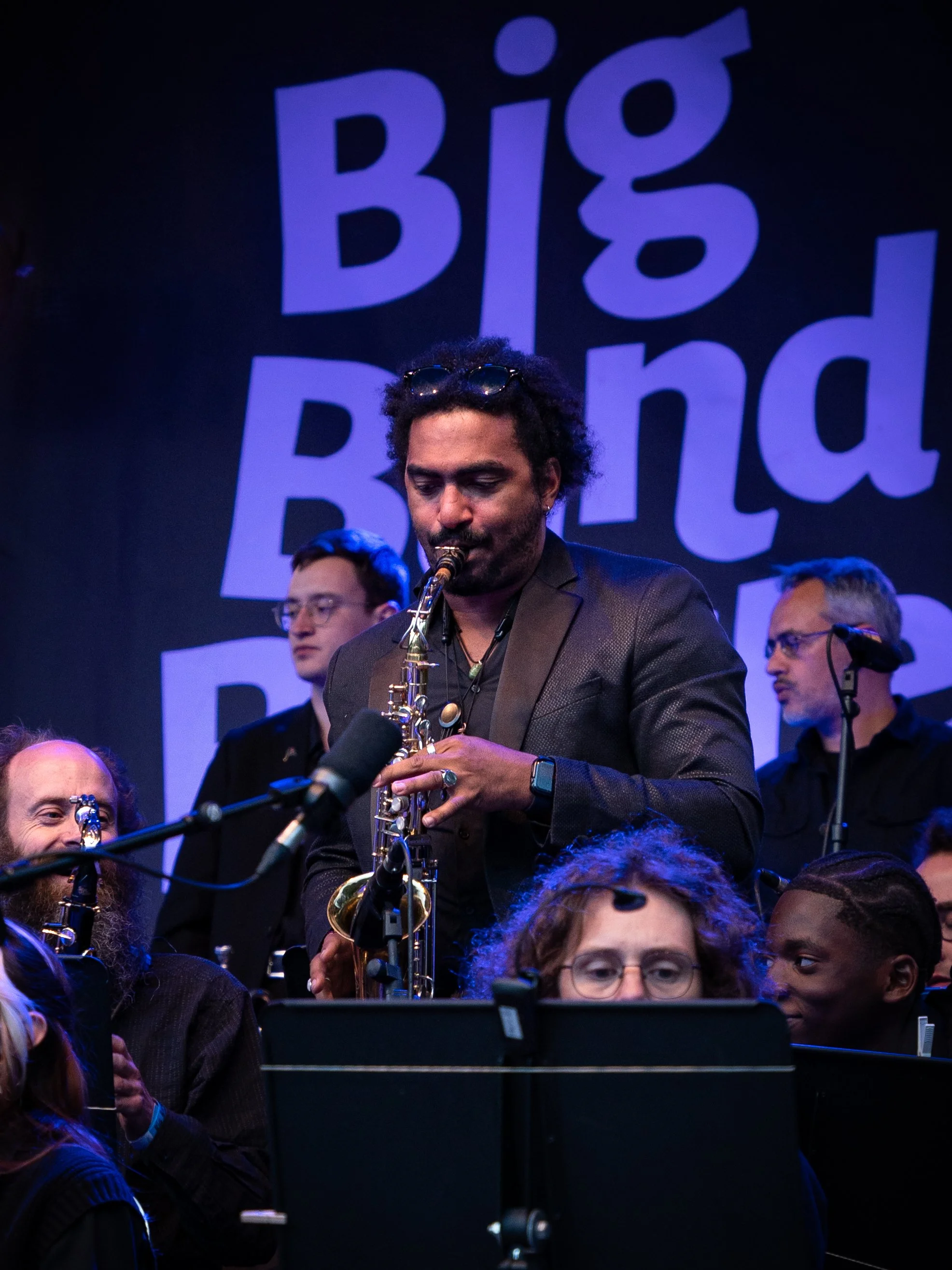 Musicien jouant du saxophone lors d'un concert avec d'autres musiciens. Un grand panneau derrière affiche le texte 'Big Bend' en lettres violettes.