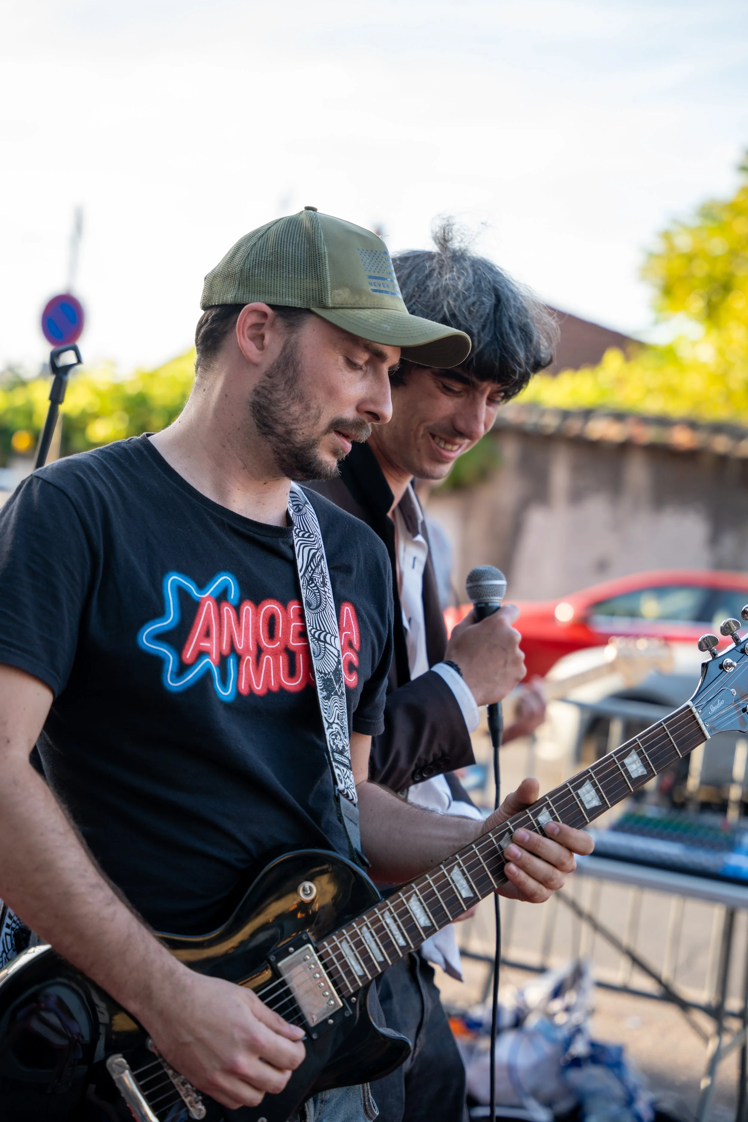 Deux hommes jouent de la guitare lors d'une performance en extérieur, avec des arbres et des voitures en arrière-plan.