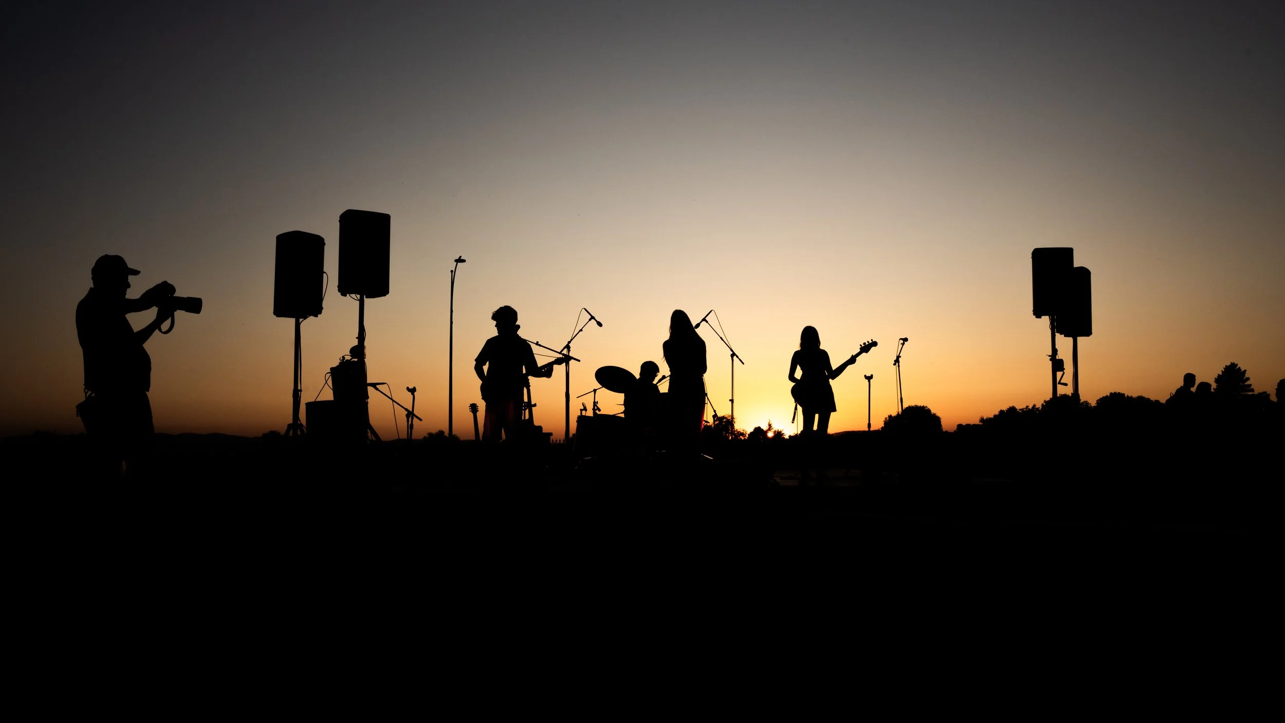 Silhouettes d'un groupe de musiciens jouant en plein air au coucher du soleil avec des haut-parleurs et du matériel audio.