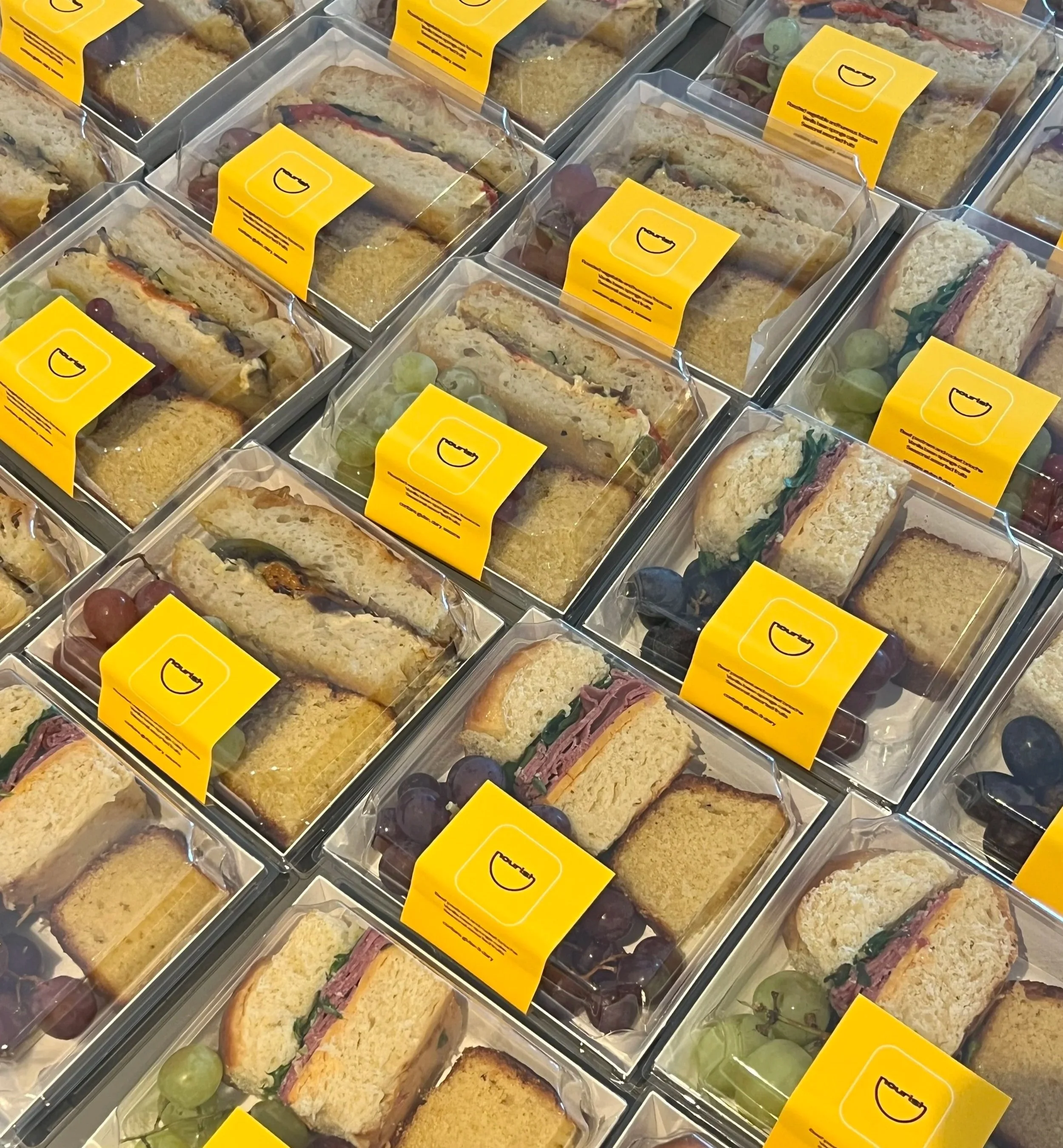 Multiple packaged sandwiches with bread, deli meats, lettuce, and tomatoes, arranged in plastic containers with yellow labels, alongside green grapes.