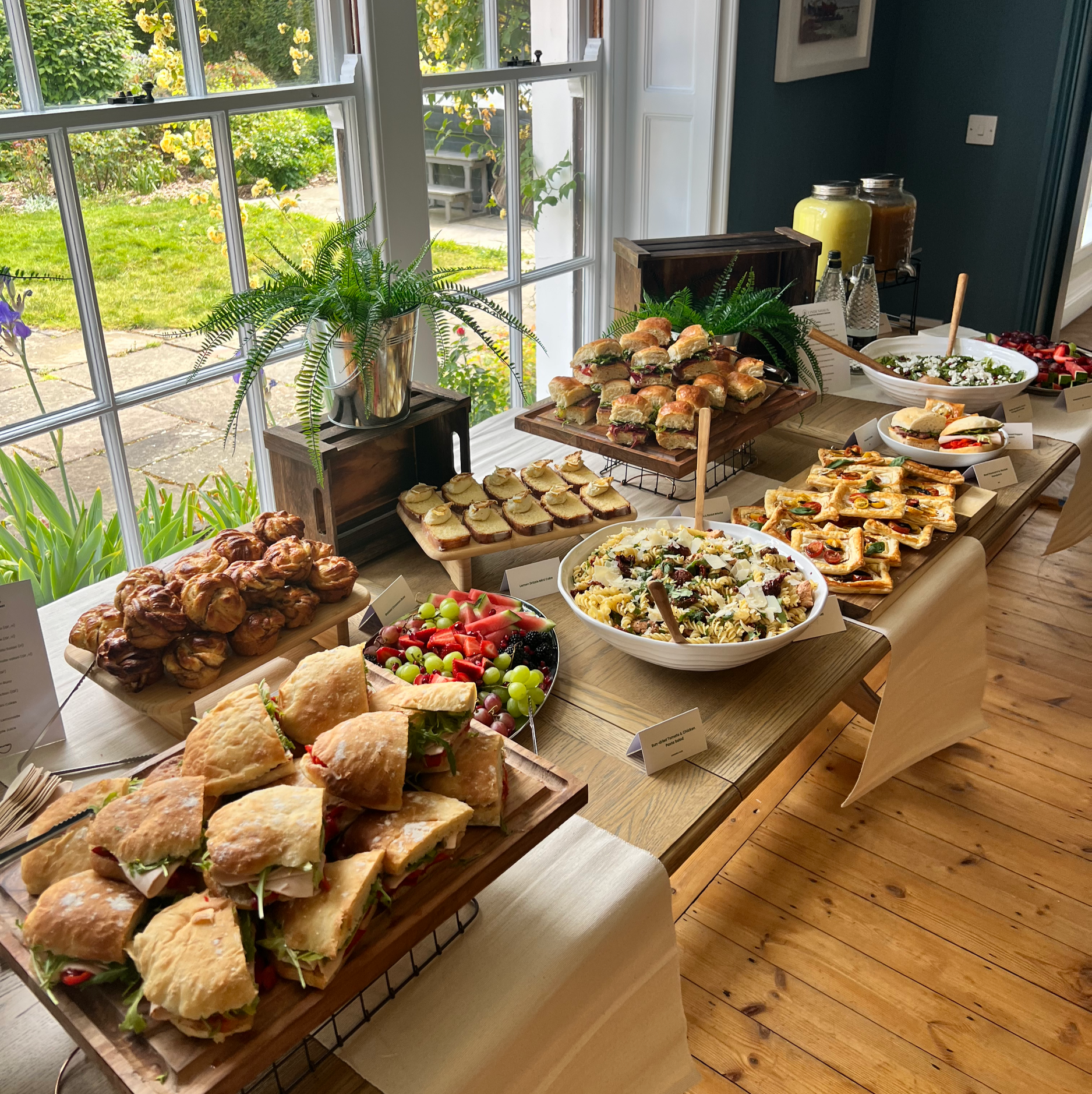 A variety of sandwiches, salads, fruit, and baked goods arranged on a table by a large window with a garden view.