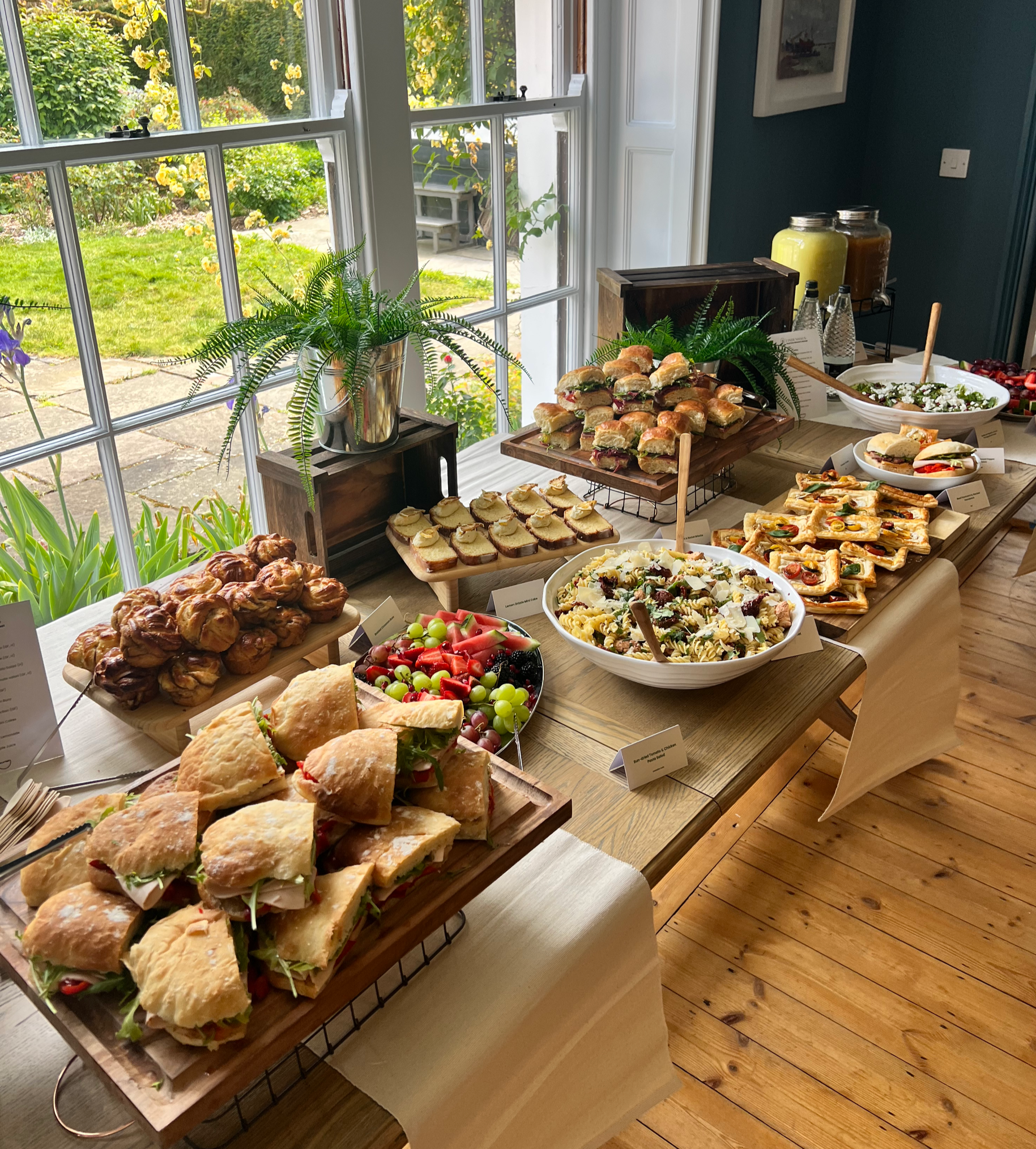 A table set with various finger foods including sandwiches, pastries, a pasta salad, a fruit platter, and salad bowls, near a window overlooking a garden.