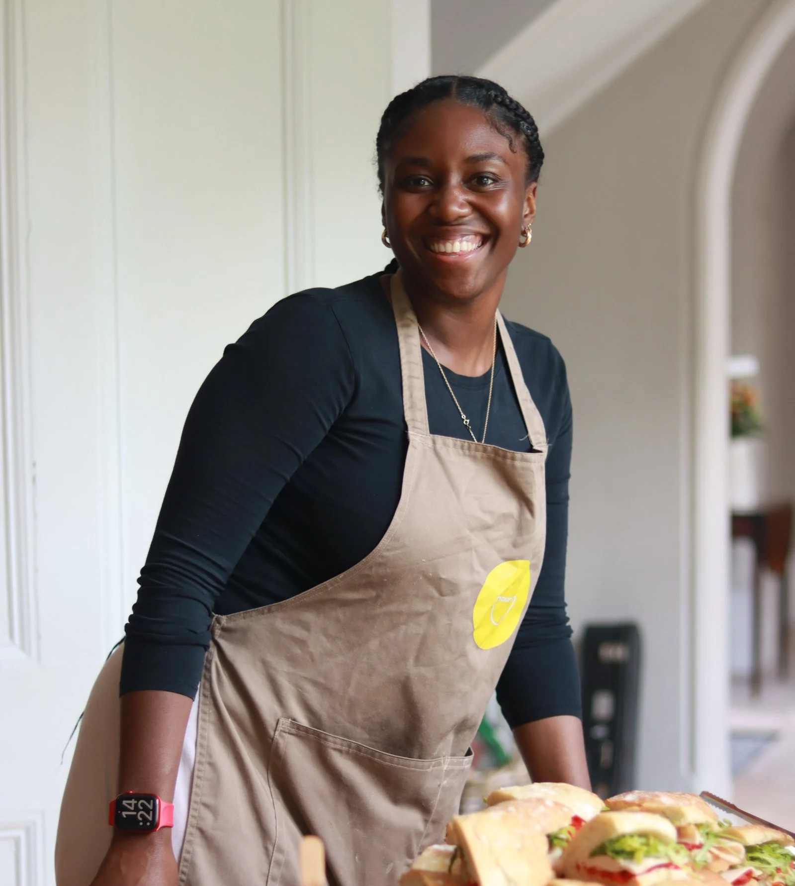 Woman smiling while preparing sandwiches in a kitchen, wearing a beige apron and a black shirt.