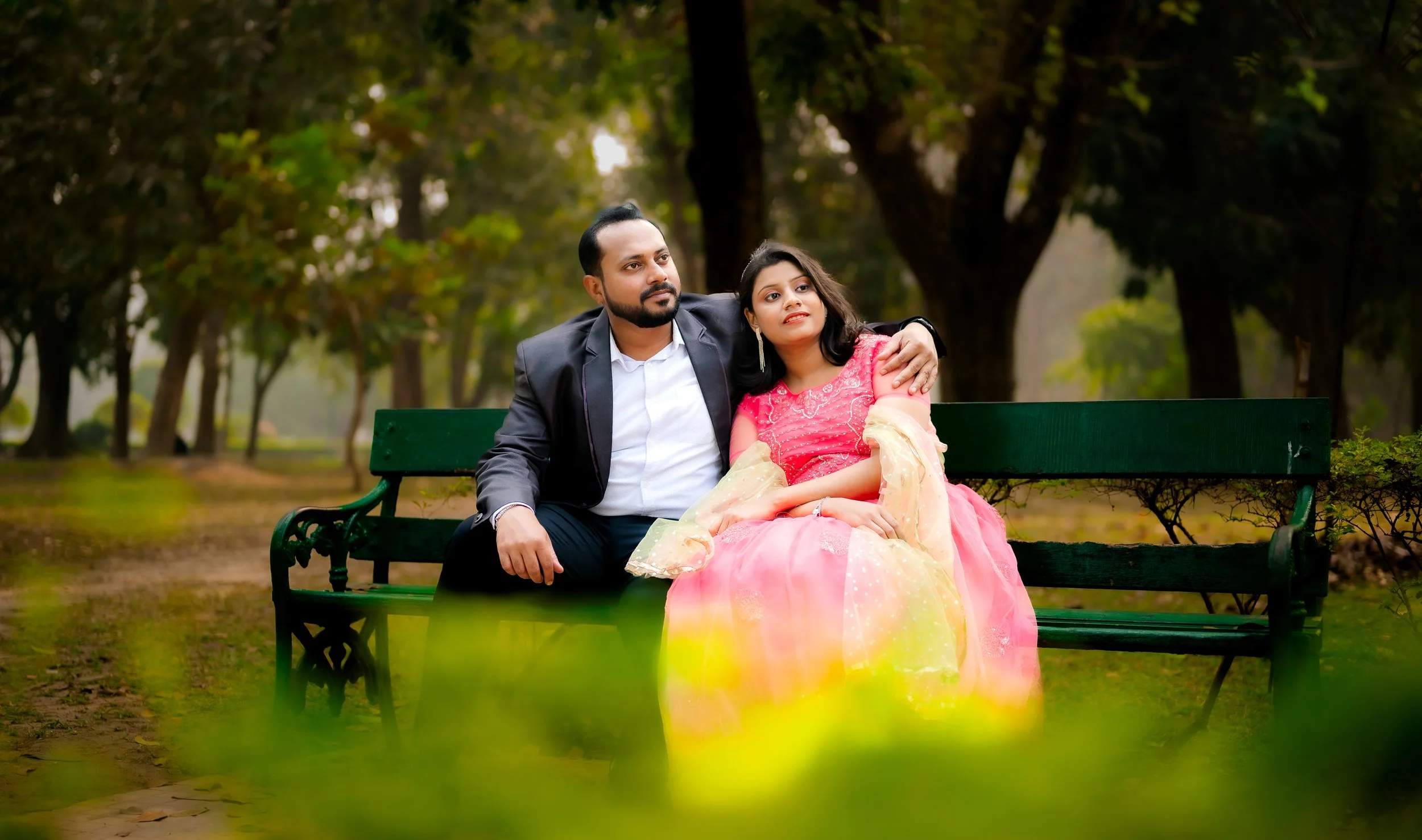 A man with a beard in a dark suit and a woman in a pink traditional dress sitting closely on a green park bench surrounded by trees.