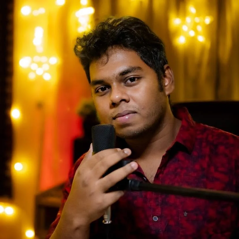Young man with dark curly hair holding a microphone, wearing a red patterned shirt, standing in front of a warmly lit background with string lights.