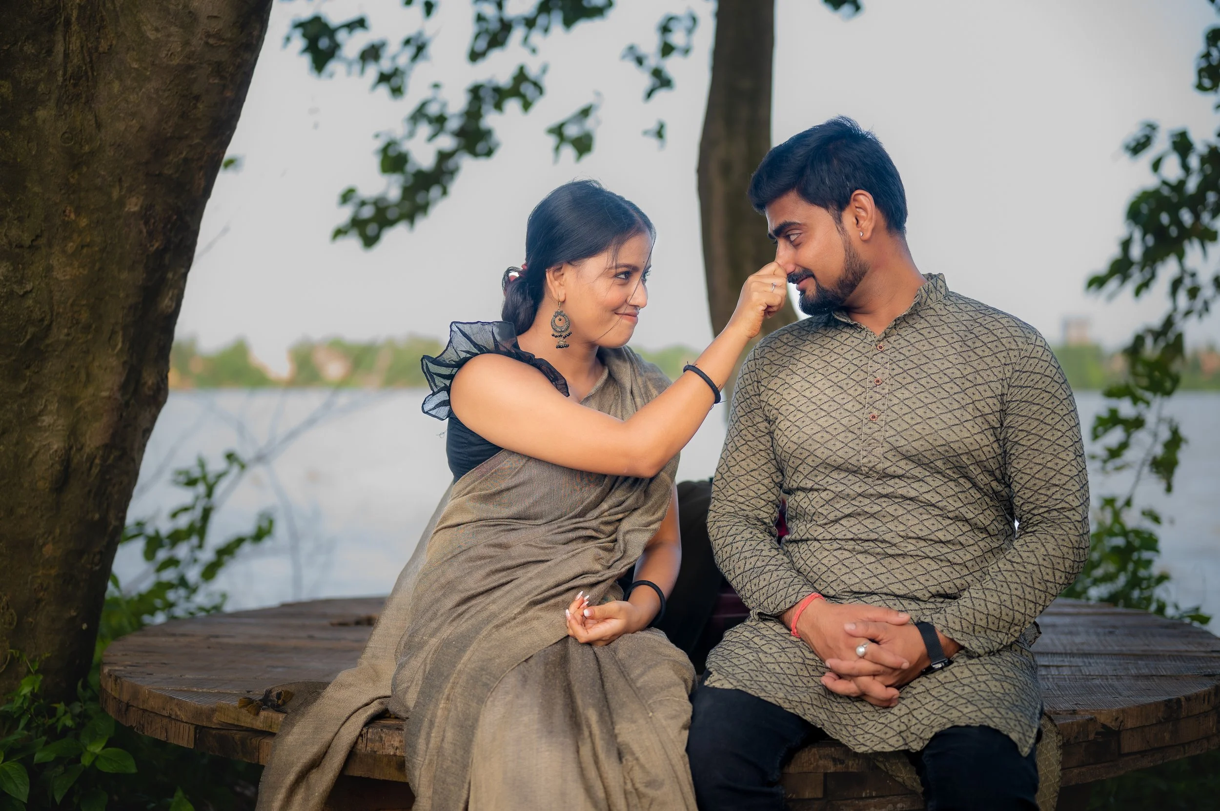 A woman touching a man's nose with her finger while sitting on a wooden platform by a tree near a body of water.