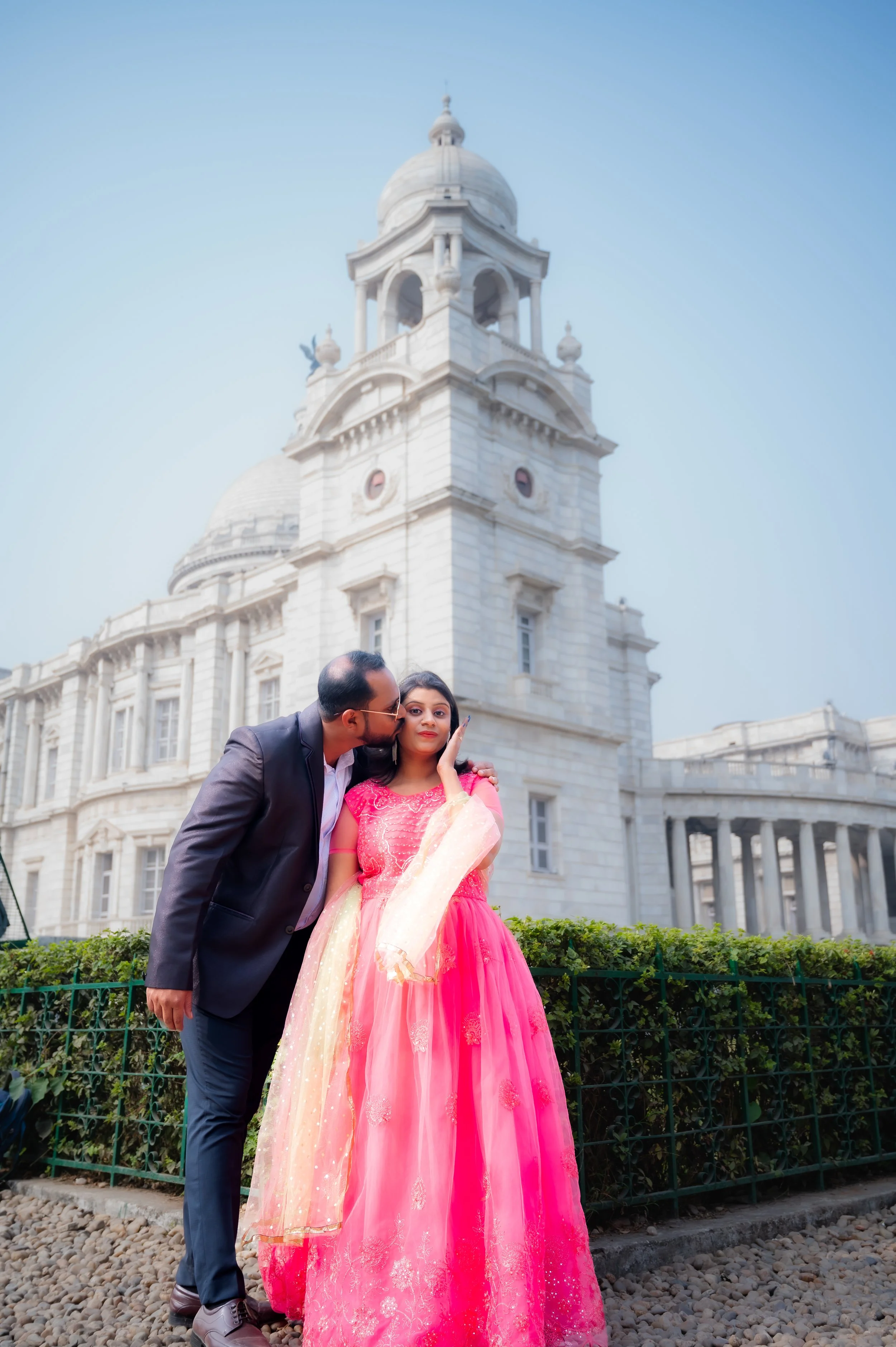 A man in a suit kisses a woman in a pink and gold traditional outfit on the cheek in front of a historic white building with a clock tower.