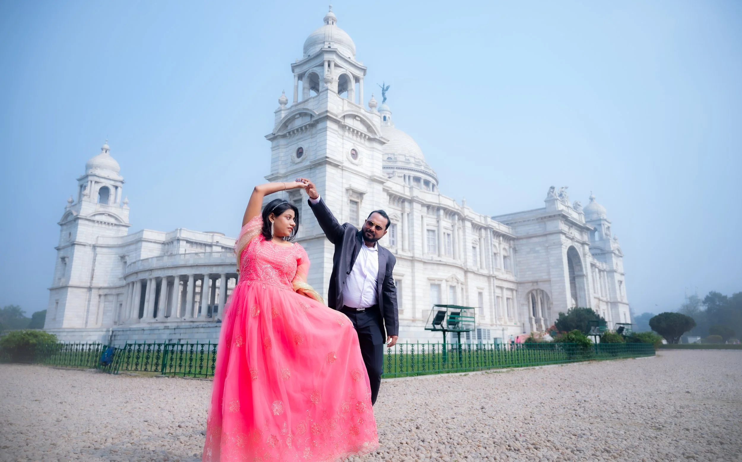 A man and a woman dancing in front of a white historic building with domes and towers, on a gravel area with a green fence and trees in the background.