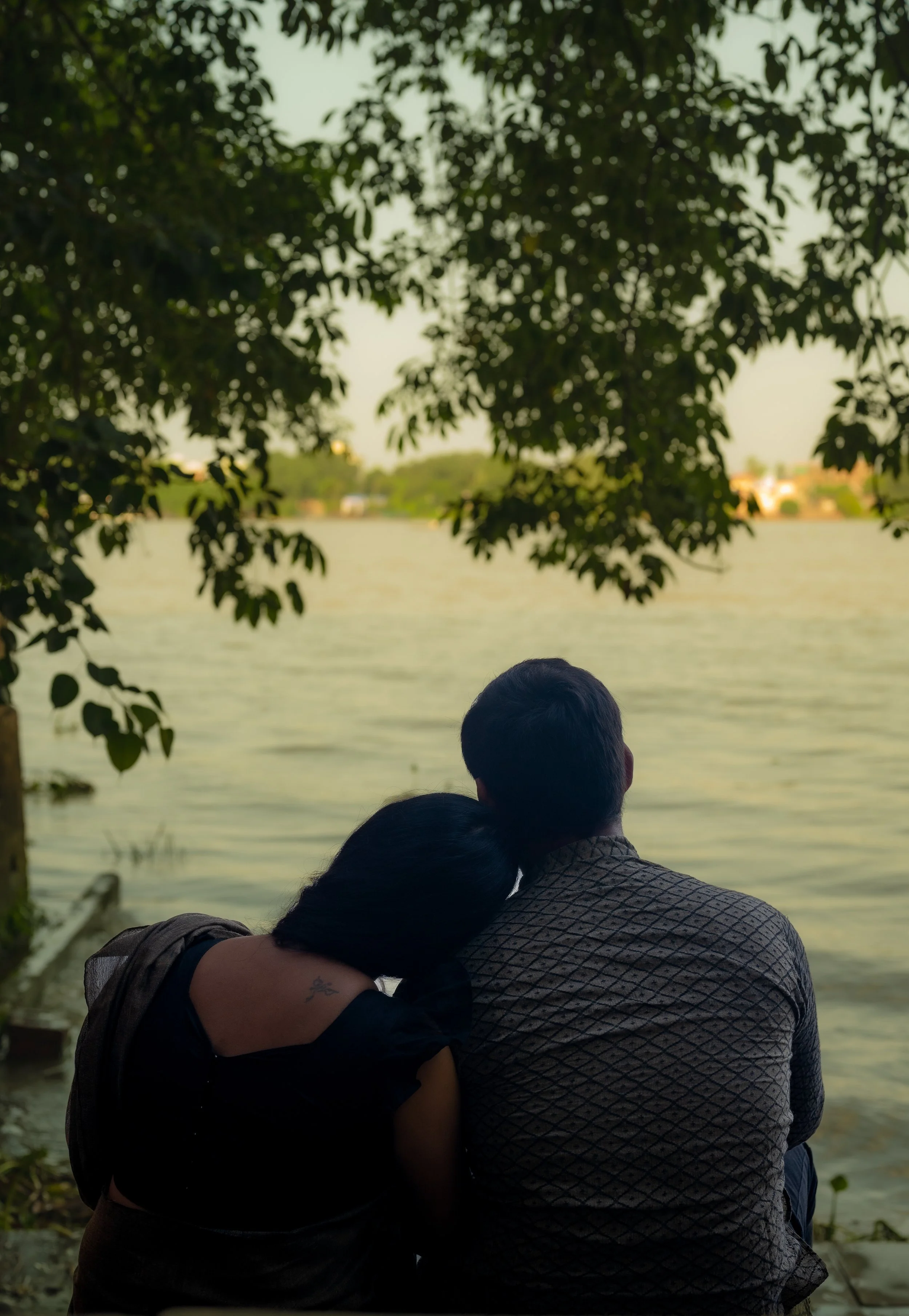 A couple sitting together by the water, embracing and looking out over a lake or river with trees and houses in the background.