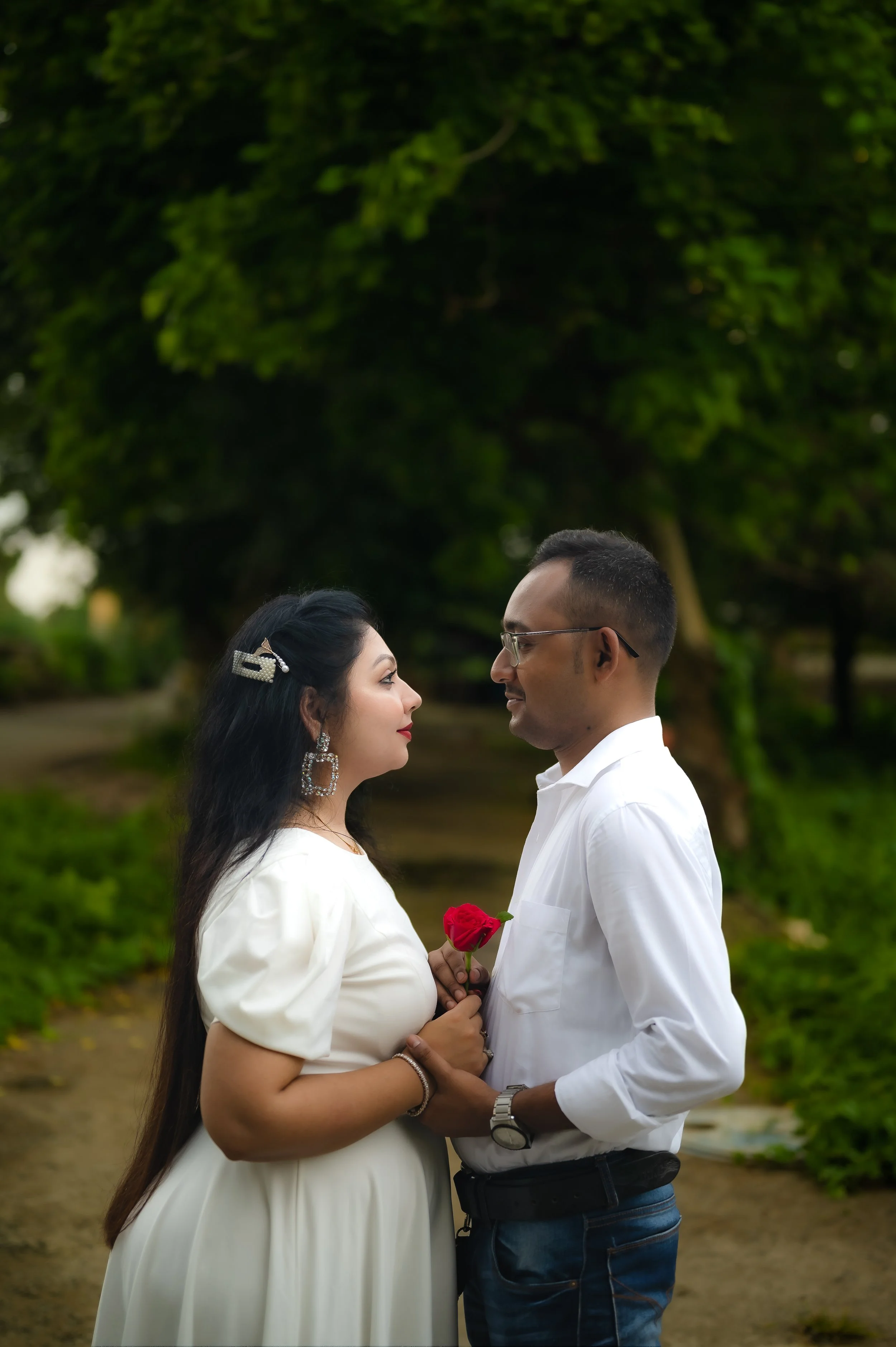 A couple stands close together outdoors, facing each other, with the woman holding a red rose. They are dressed in white, and the scene is surrounded by greenery.