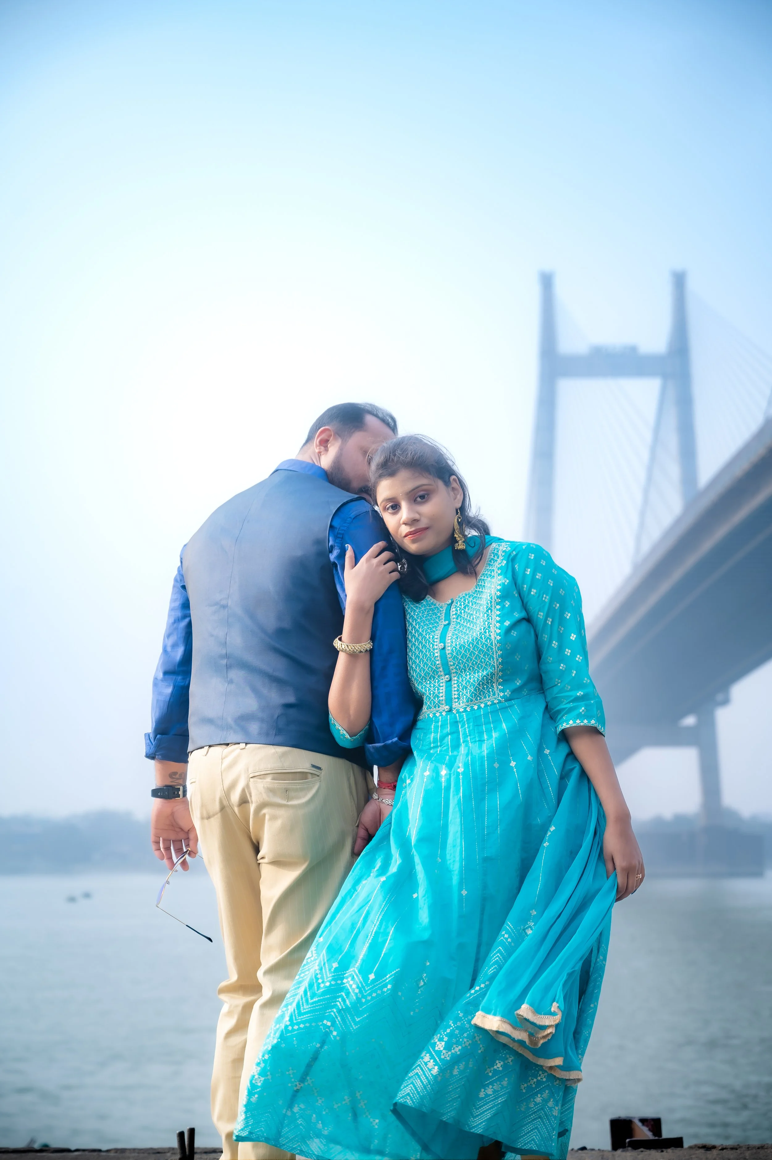 A man and woman standing close together outdoors near a body of water, with a large bridge in the background. The woman is wearing a blue traditional dress, and the man is dressed in a blue vest and beige trousers.