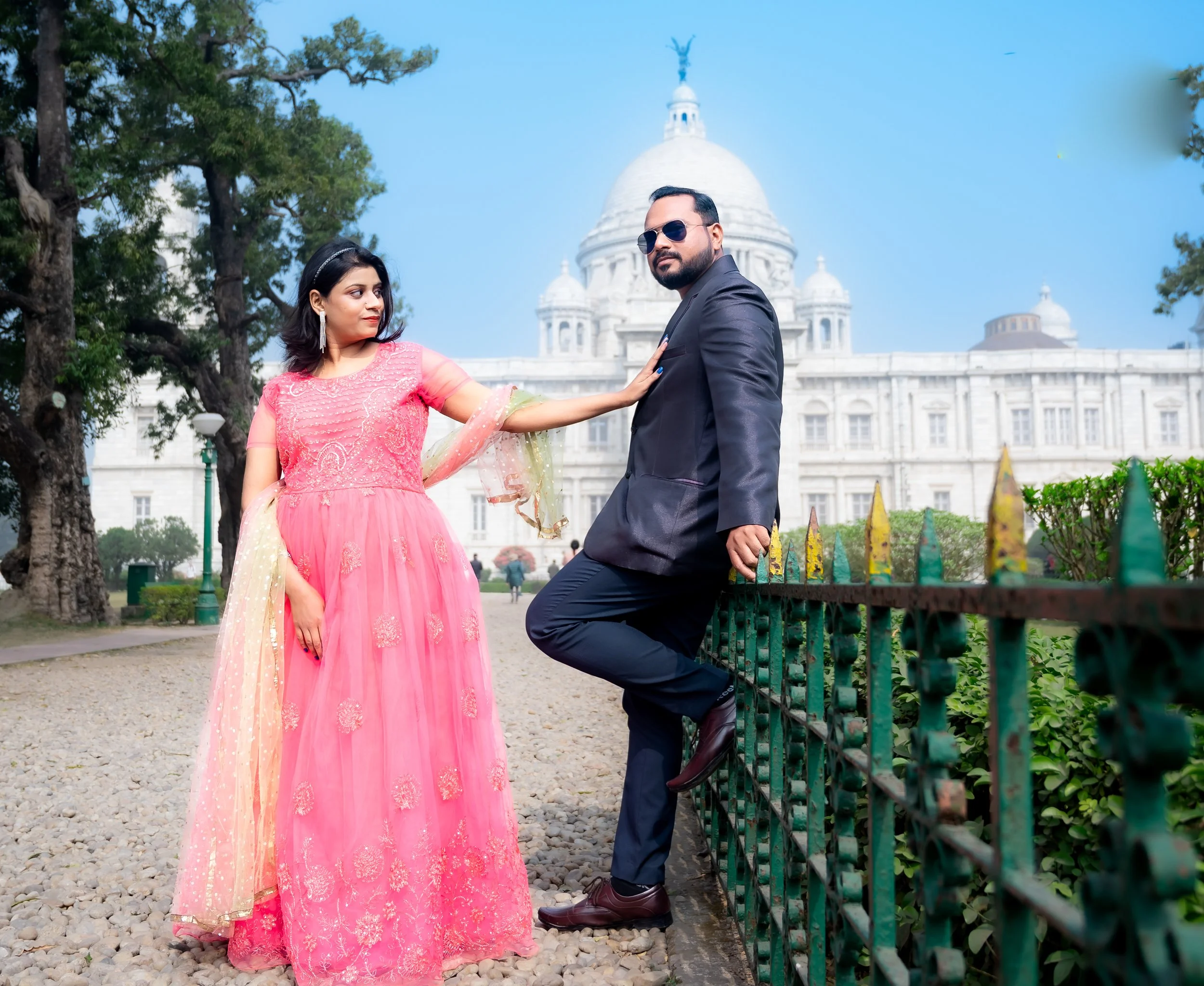 A man in a black suit and sunglasses leaning against a green fence with one foot on a rail, and a woman in a pink traditional dress gently touching his chest in front of a white building with a dome and statues, with trees surrounding them under a blue sky.