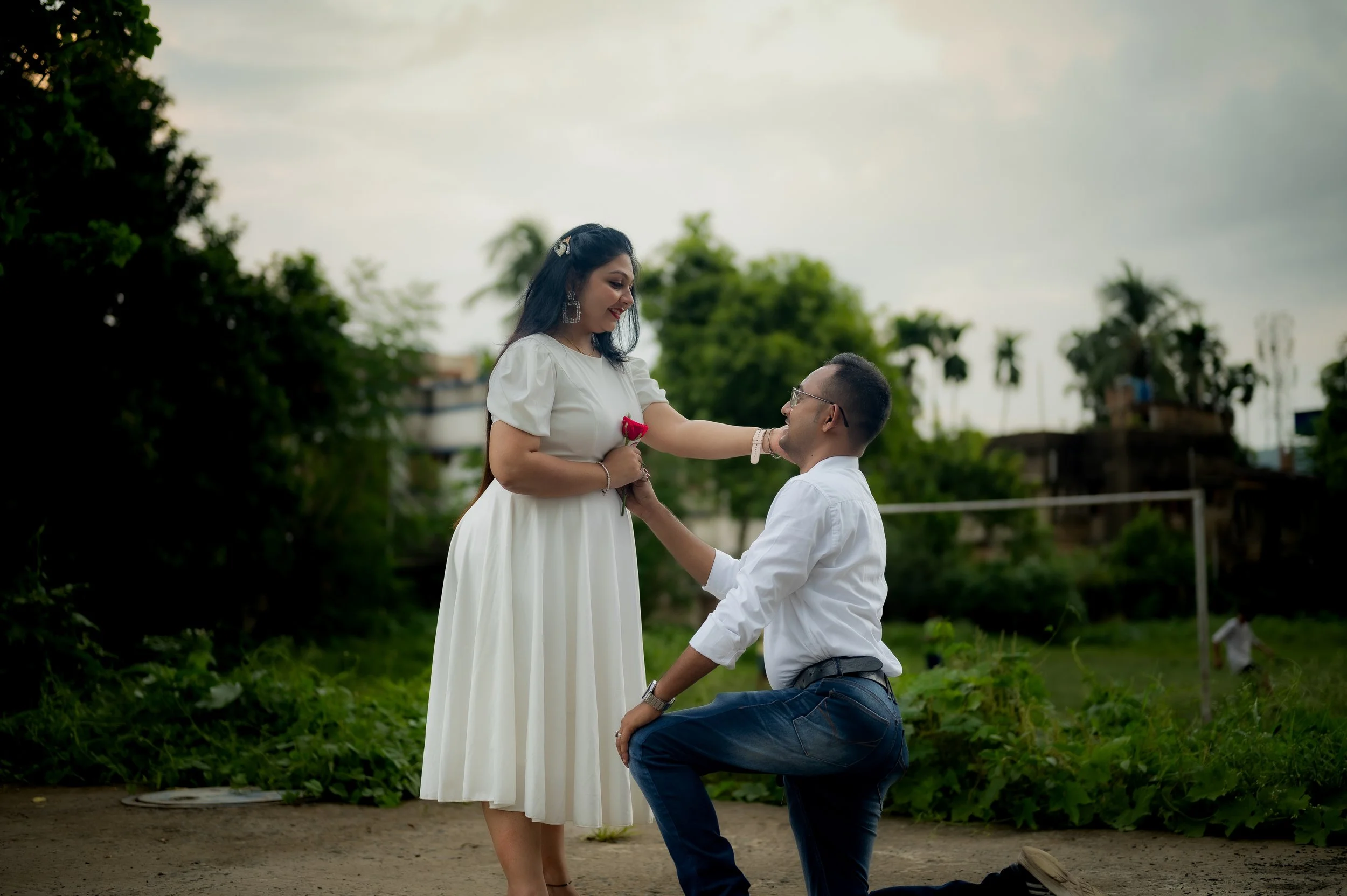 A man on one knee proposing to a woman in an outdoor park, she is holding a red rose and smiling, surrounded by trees and cloudy sky.
