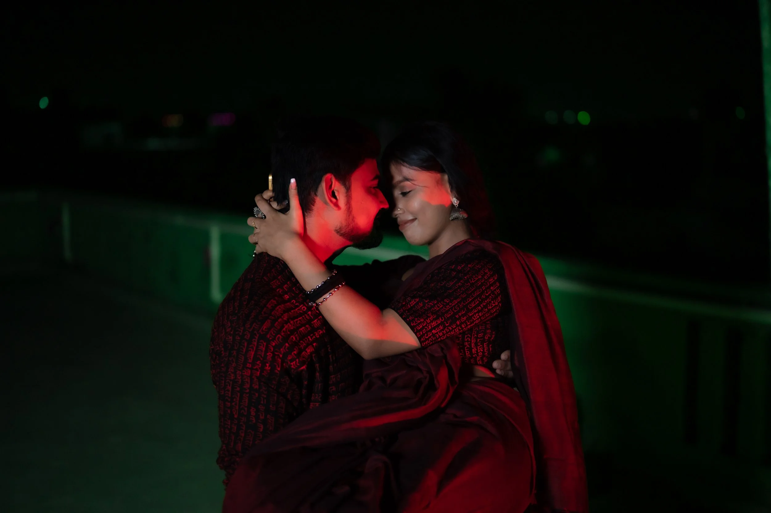 A man and woman passionately embrace each other on a rooftop at night with city lights in the background, illuminated by red and green lighting.