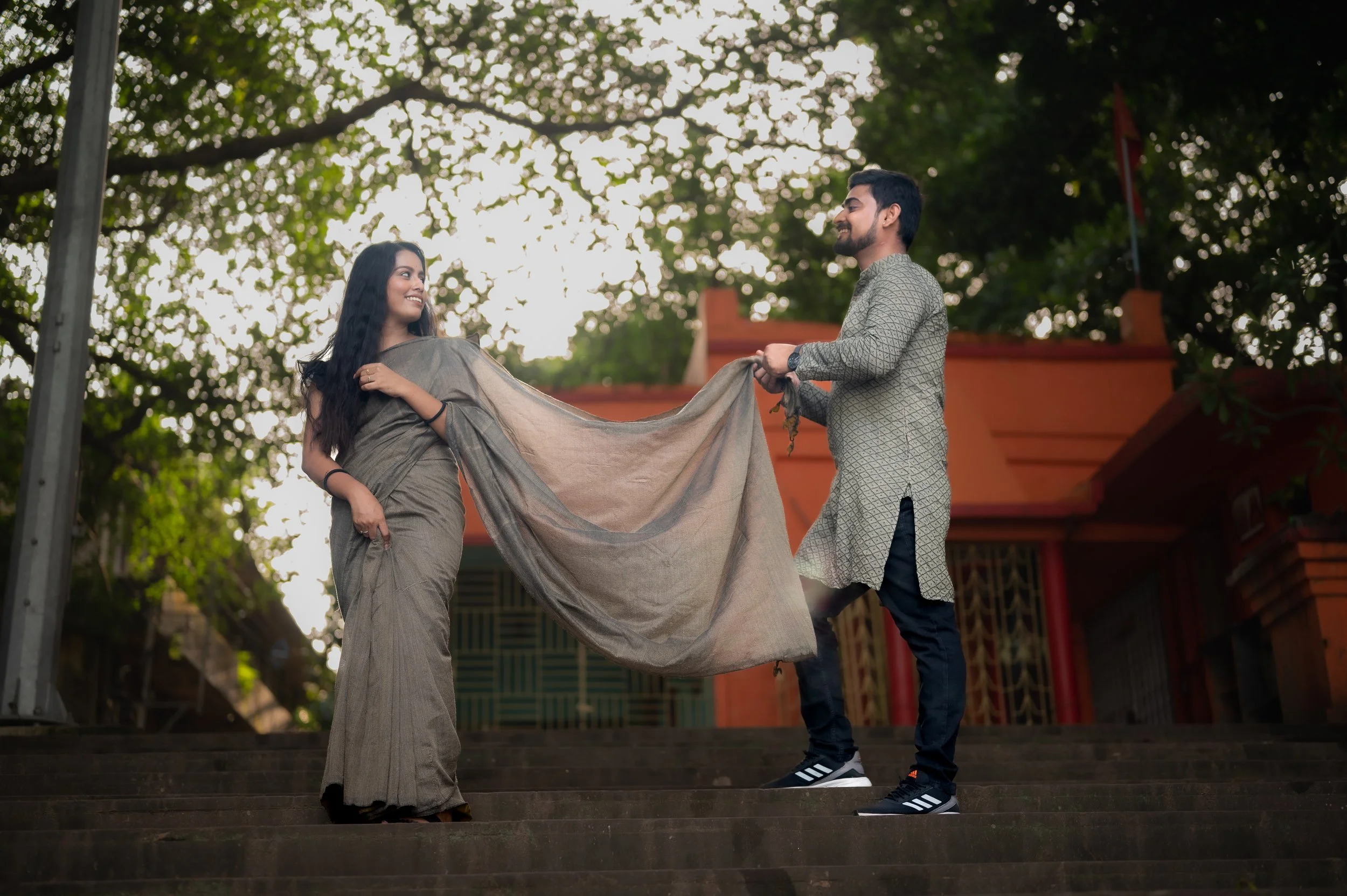 A man and woman standing on steps outdoors, exchanging a cloth, with trees and a building in the background. The woman is wearing traditional attire, and the man is casually dressed.