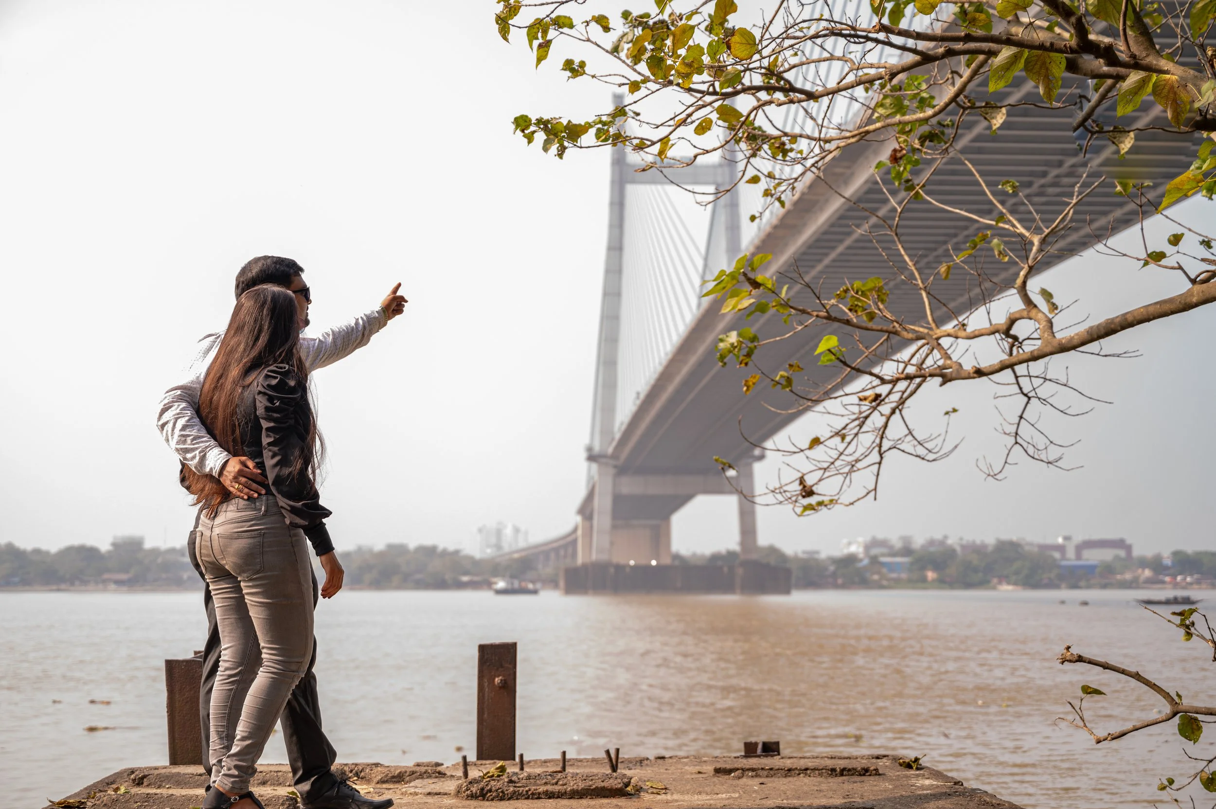 Two women standing by the riverbank under a tree, with a large bridge in the background. One woman is pointing towards the bridge.