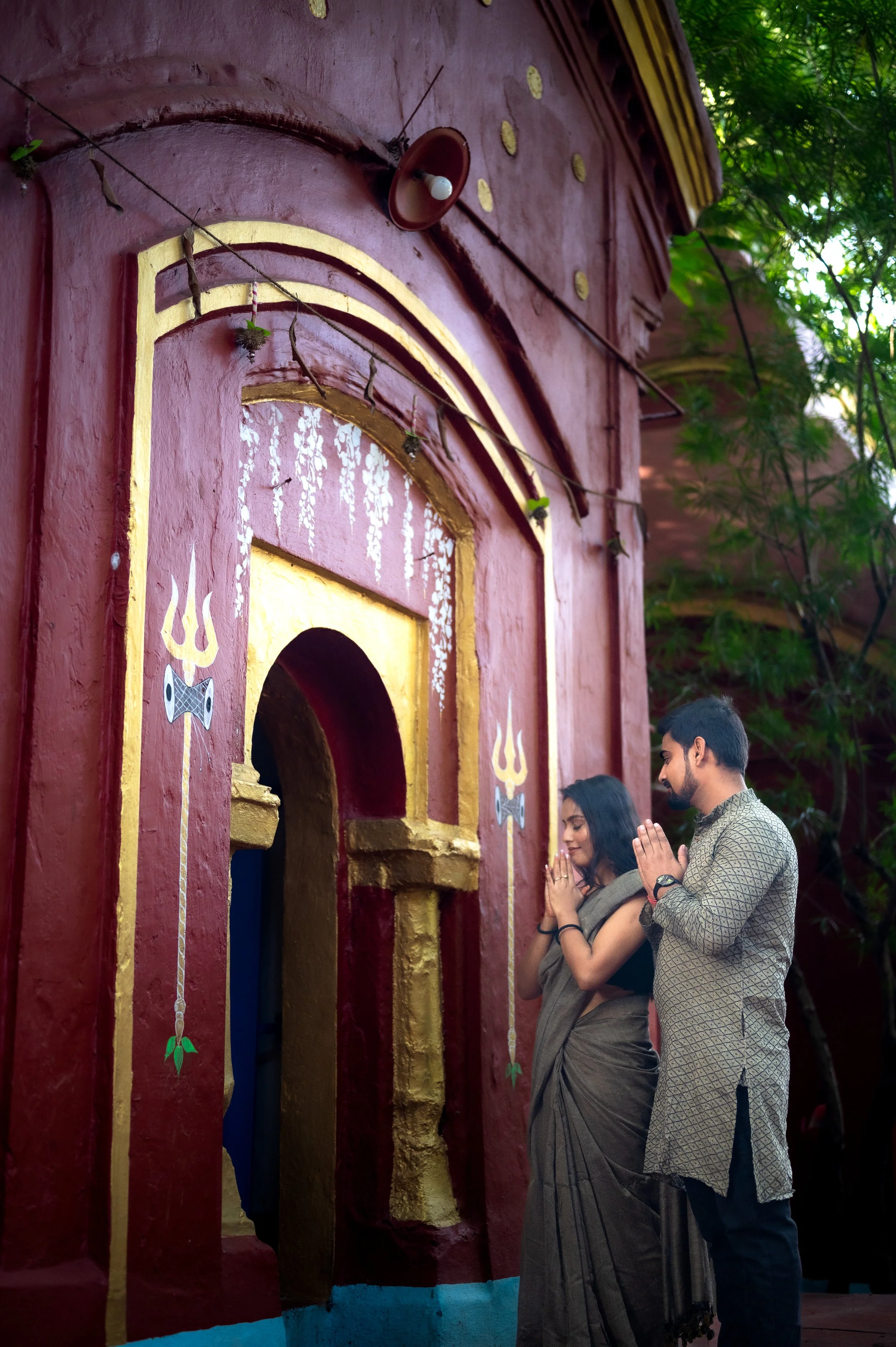 Two people praying in front of a painted religious shrine, with their hands pressed together and their heads bowed. The shrine is decorated with gold accents and symbols, set against a red wall with green foliage in the background.