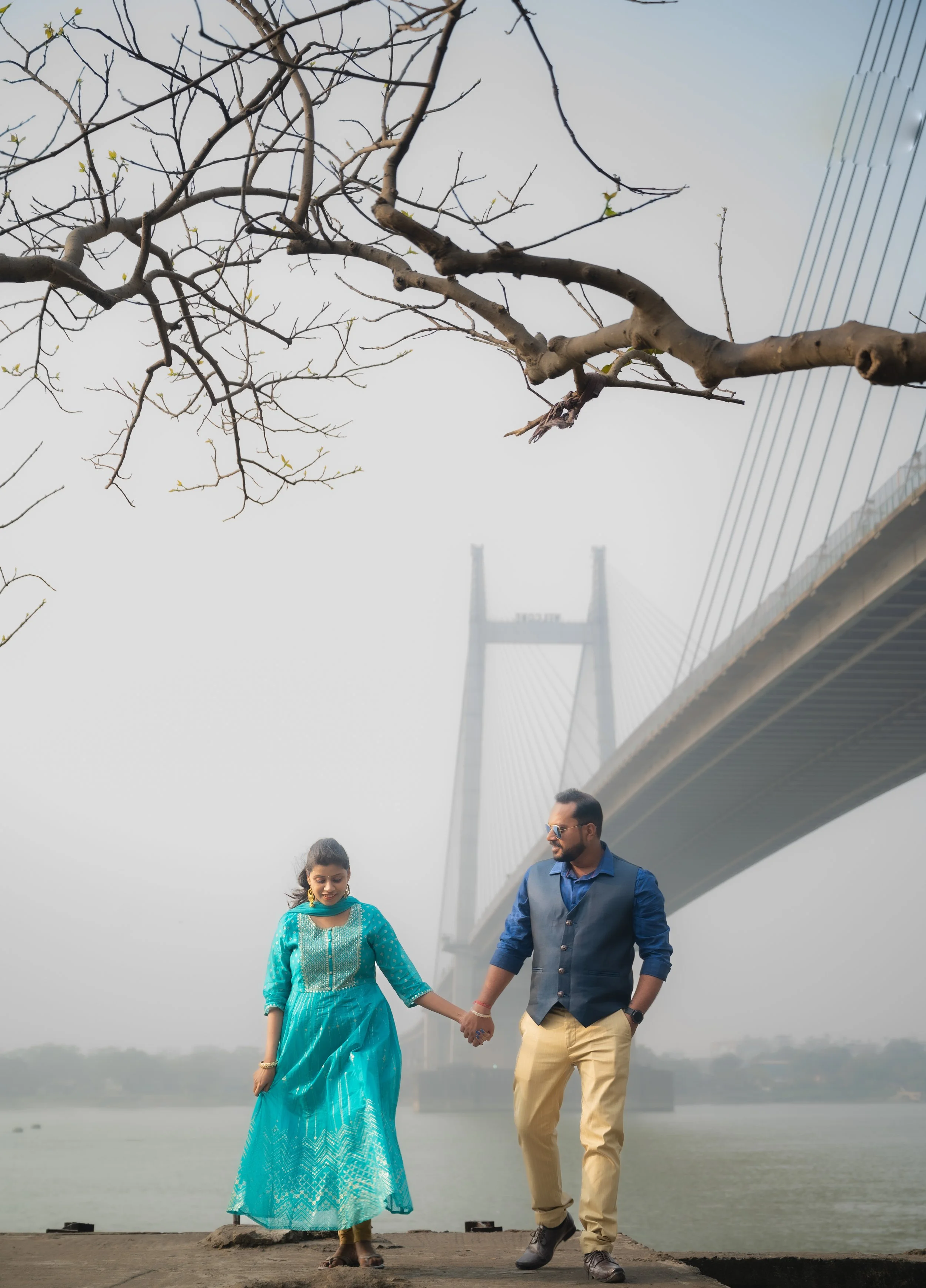 A couple walking hand in hand near a river with a bridge in the background, and a tree with budding leaves. The woman is wearing a turquoise dress, and the man is wearing a blue shirt and beige pants.