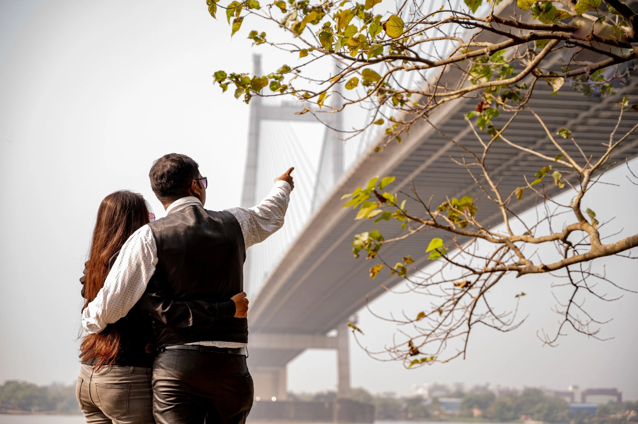 Two people standing outdoors near a tree with a bridge in the background, one person is pointing towards the bridge.