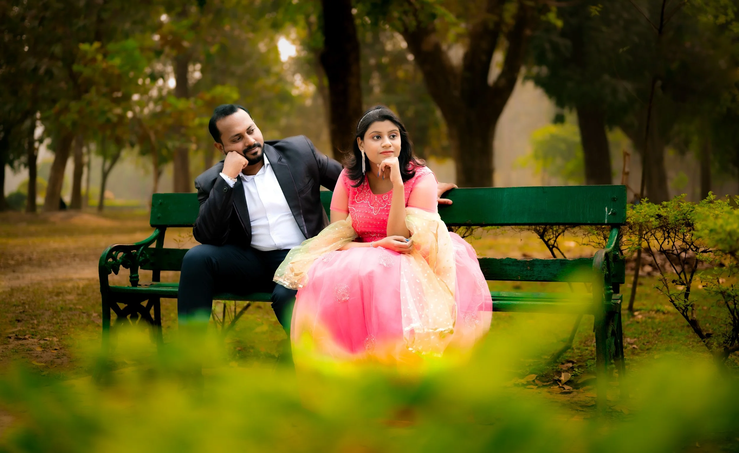 A man in a black suit and a woman in a pink and beige dress sitting on a green park bench surrounded by trees.