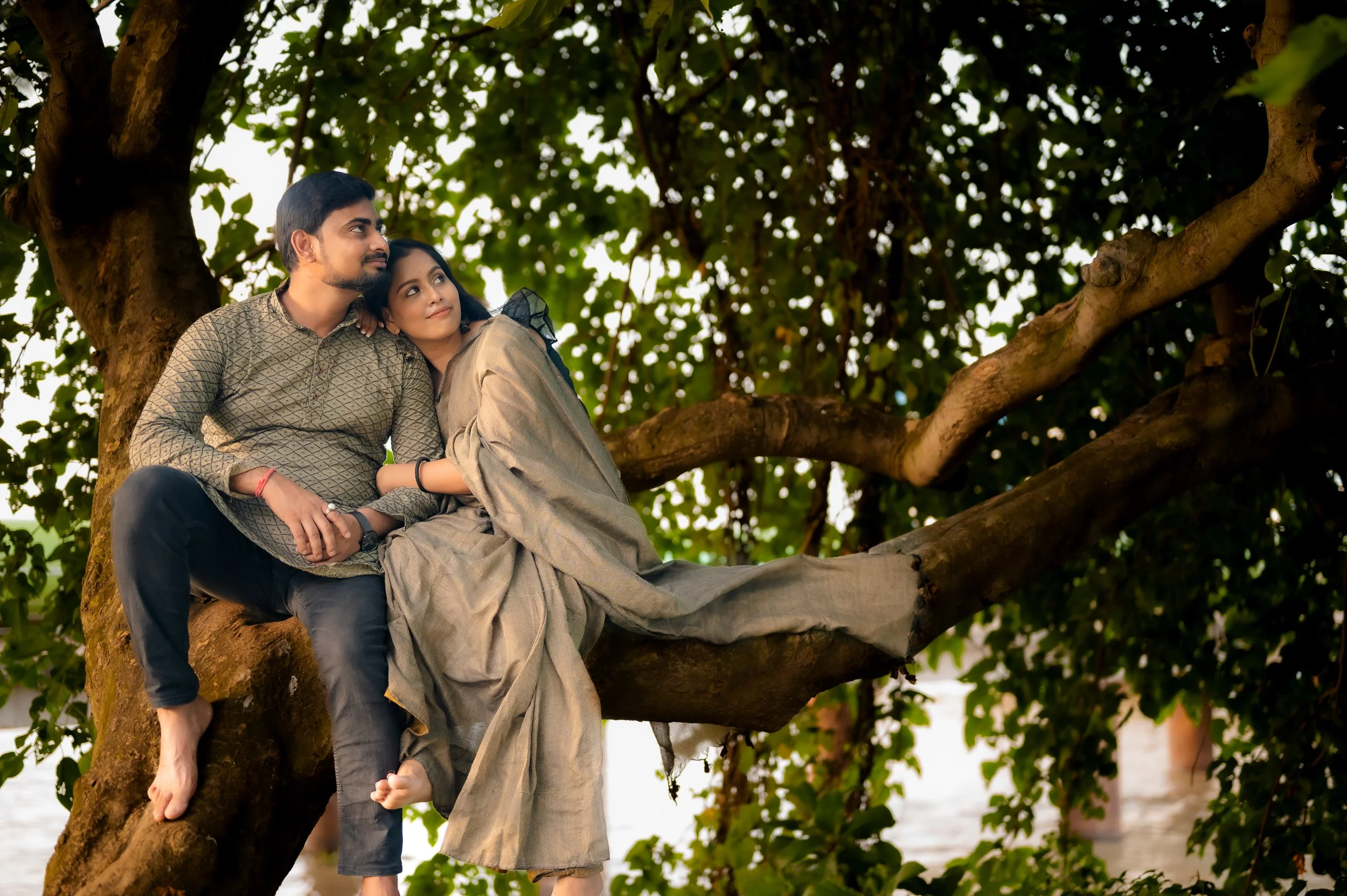 A young man and woman sitting on a large tree branch, gazing into the distance with a scenic water view in the background, surrounded by green foliage.