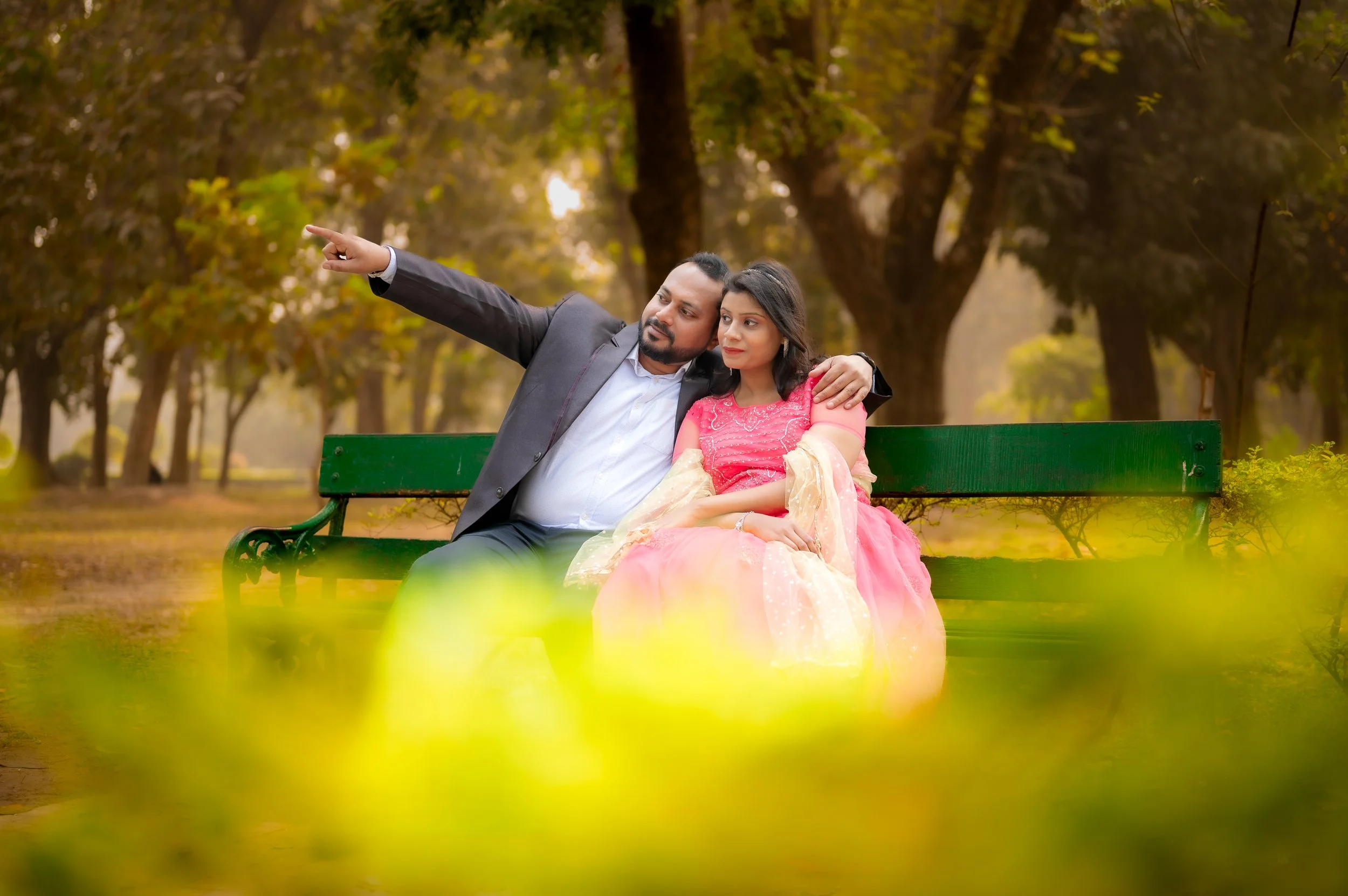 A man and woman sitting on a park bench with trees and foliage in the background. The man is pointing at something in the distance while the woman looks on. The woman is wearing a colorful traditional dress, and the man is dressed in a suit.