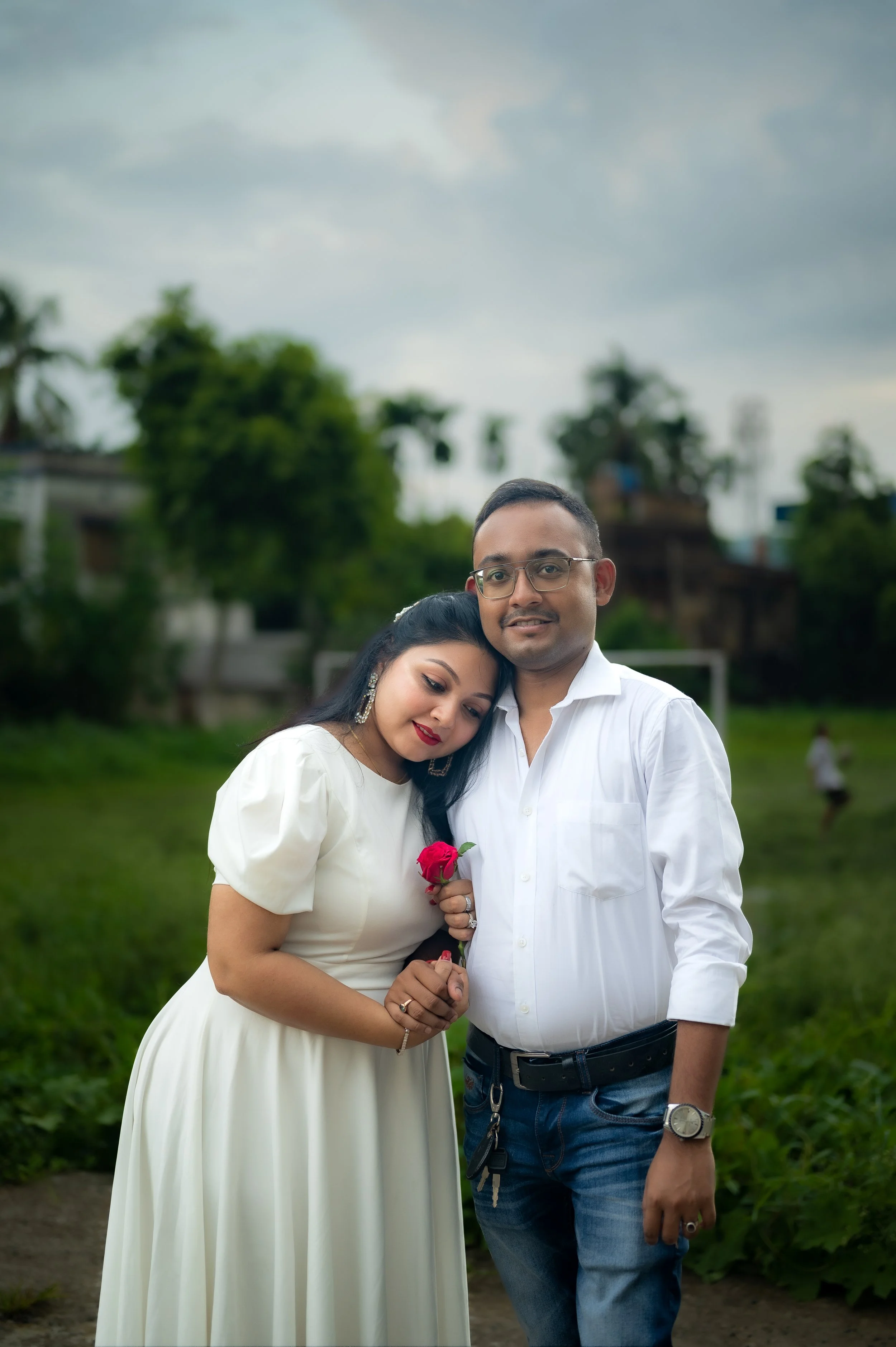 A couple standing closely together outdoors on a cloudy day, the woman holding a red rose and resting her head on the man's shoulder.