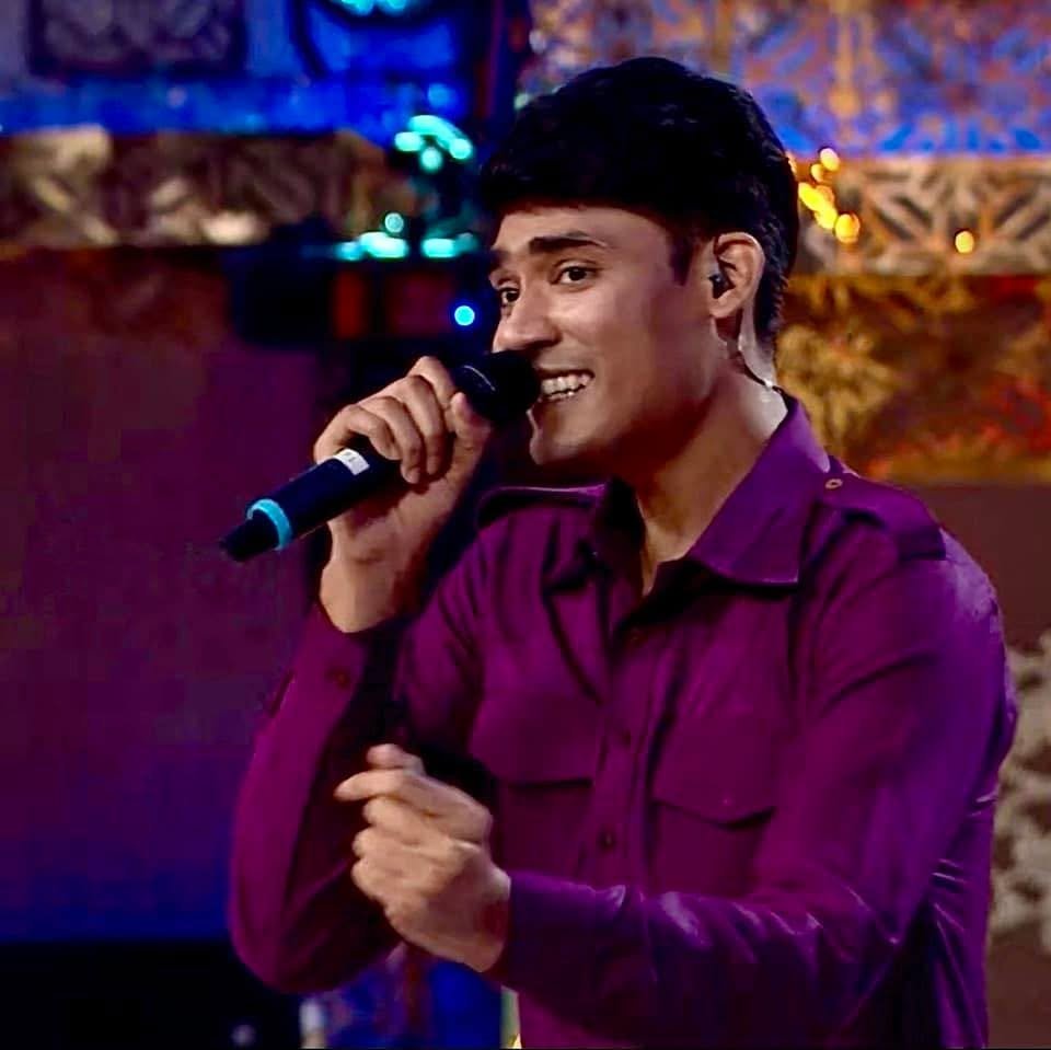 Man with black hair and a purple shirt holding a microphone, appears to be singing or speaking on stage with colorful blurred background lights.
