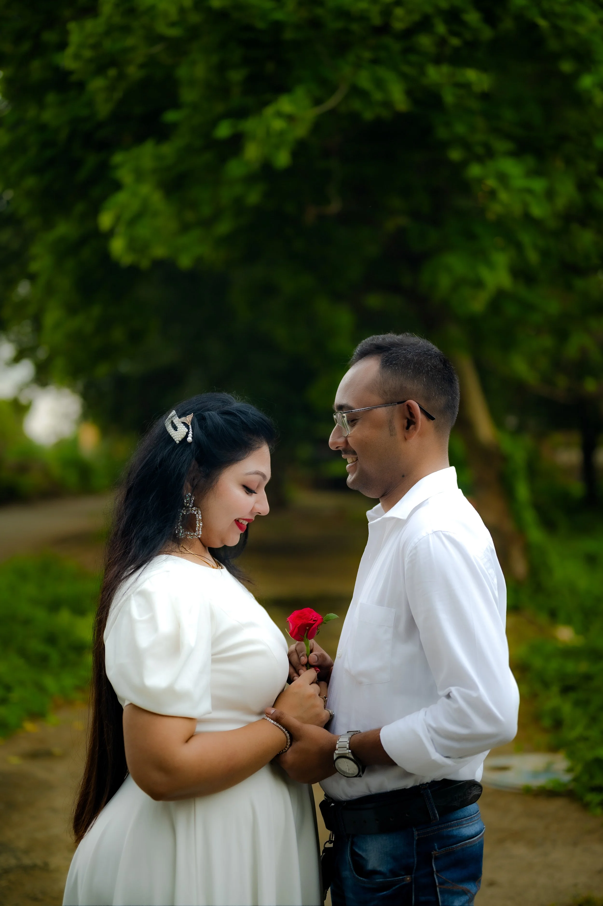 A couple standing close together outdoors, with the woman holding a red rose, embracing and smiling at each other, against a background of green trees and a dirt path.