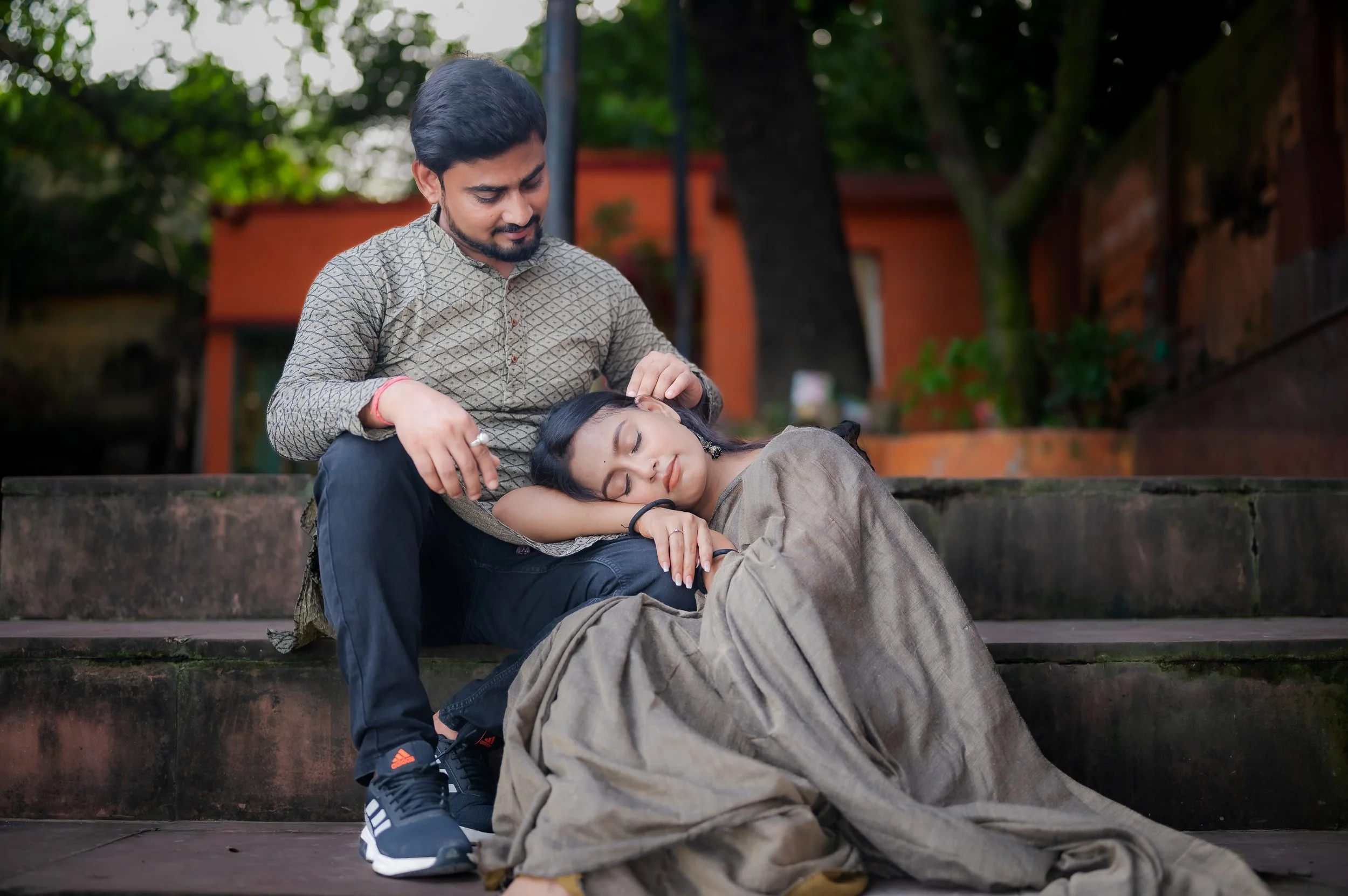 A man and a woman sitting on outdoor steps; the woman is resting her head on the man's lap while he gently strokes her hair, both with peaceful expressions, surrounded by trees and buildings.